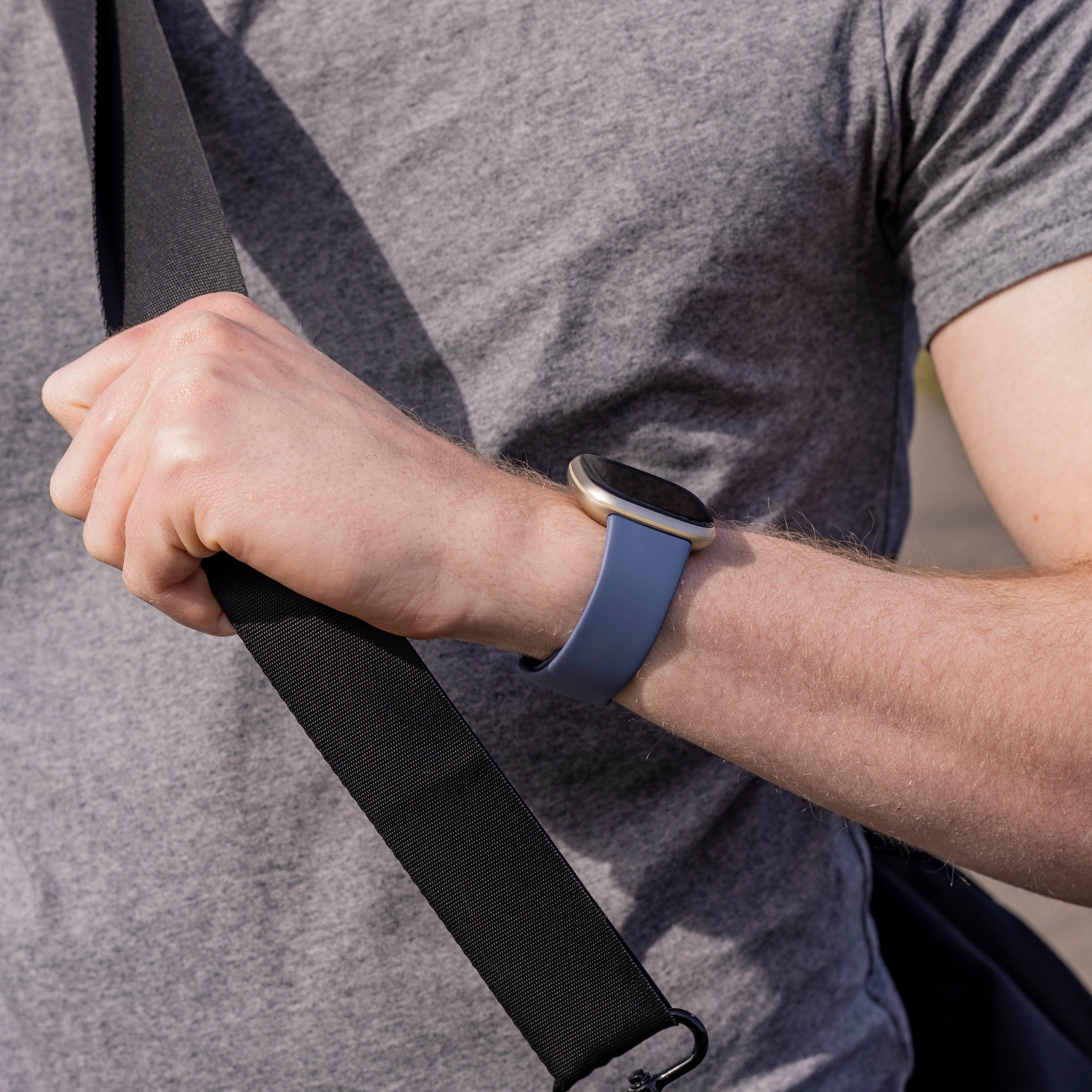 Der Arm einer Person mit einer blauen Smartwatch und einem schwarzen Taschenriemen. Die Person trägt ein graues Hemd.
