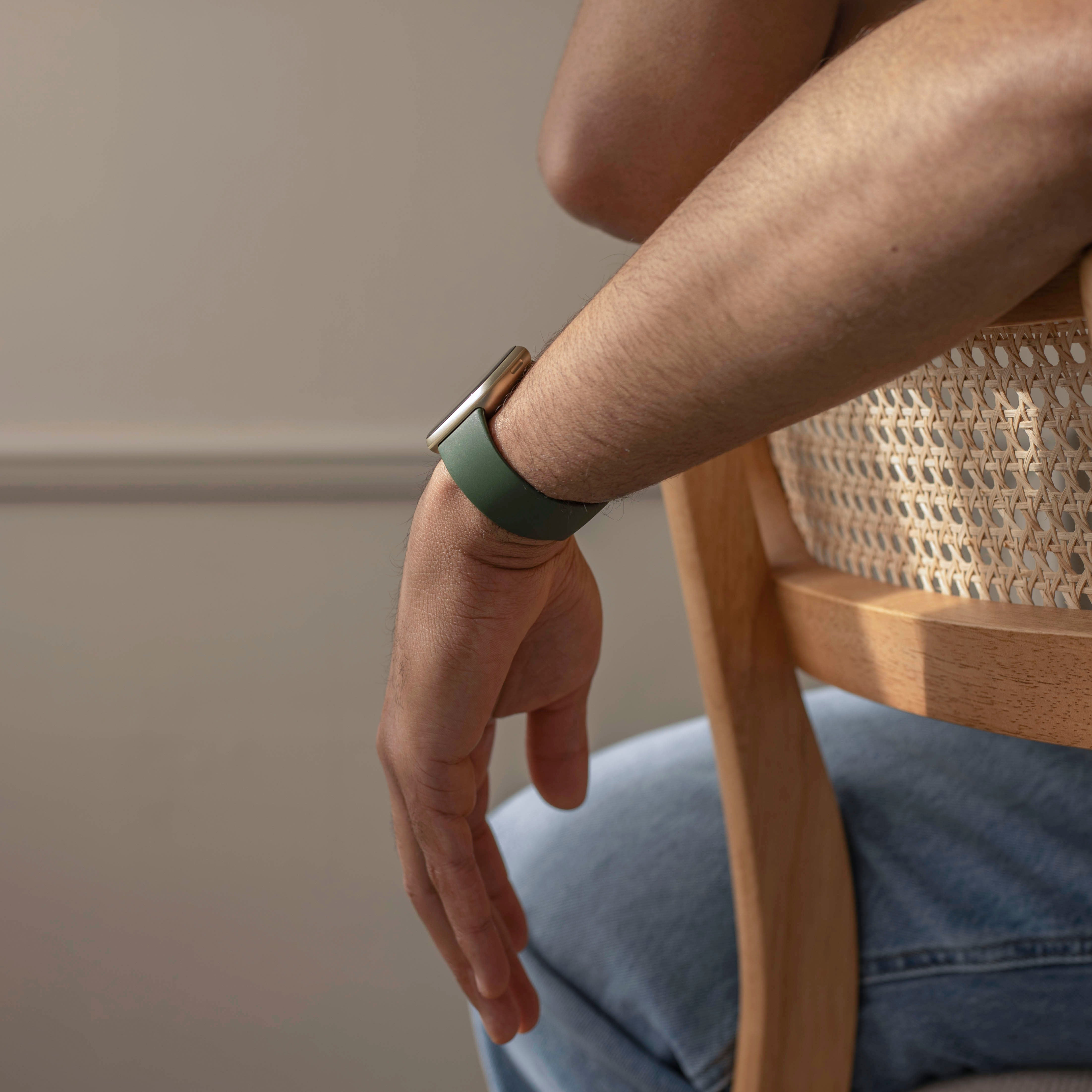 Der Arm einer Person mit einer grünen Uhr ruht auf der Lehne eines Korbstuhls. Die Person trägt blaue Jeans.