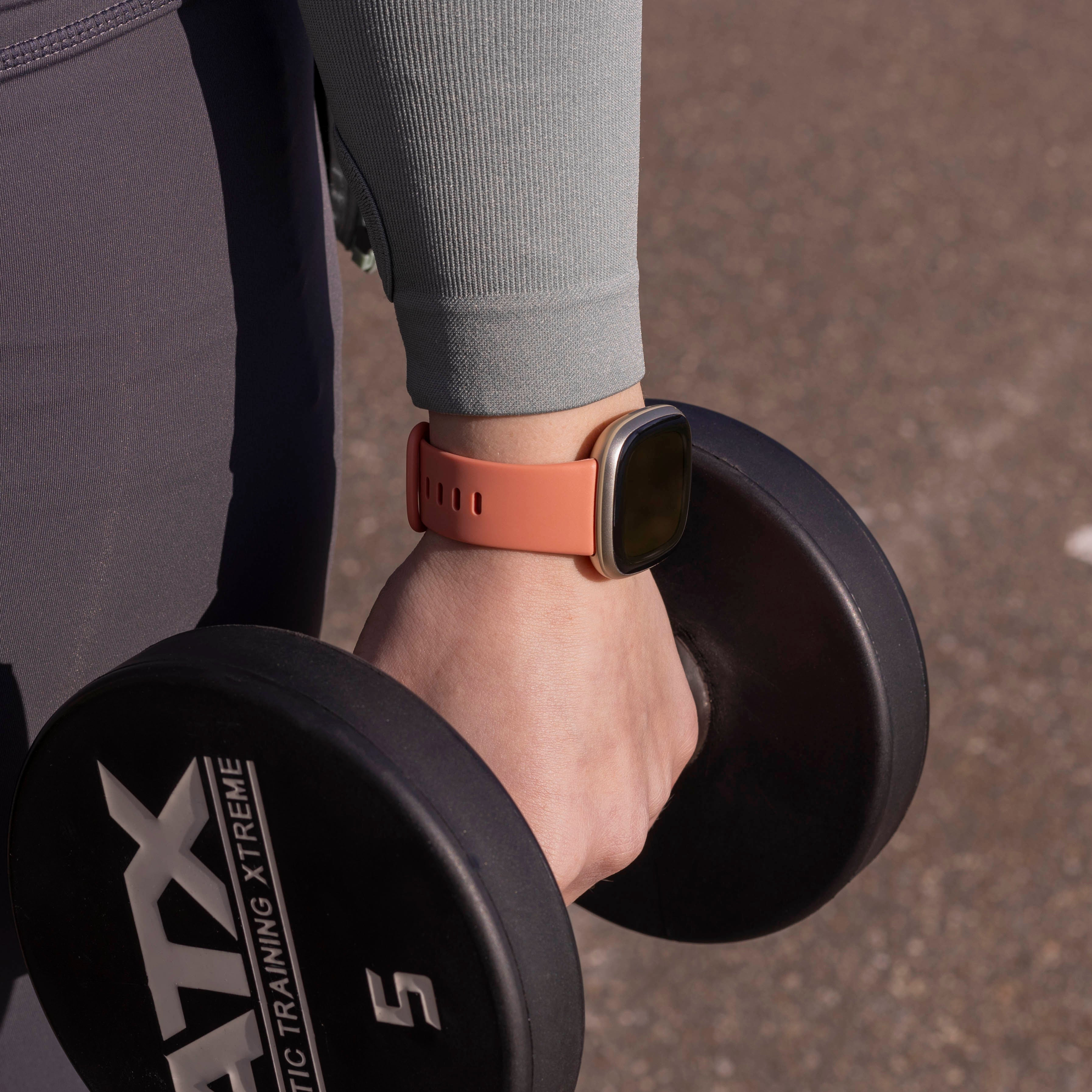 Person hält eine Hantel und trägt eine Uhr. Die Uhr hat ein orangefarbenes Armband. Die Person trägt graue Hosen und ein grünes Langarmshirt.