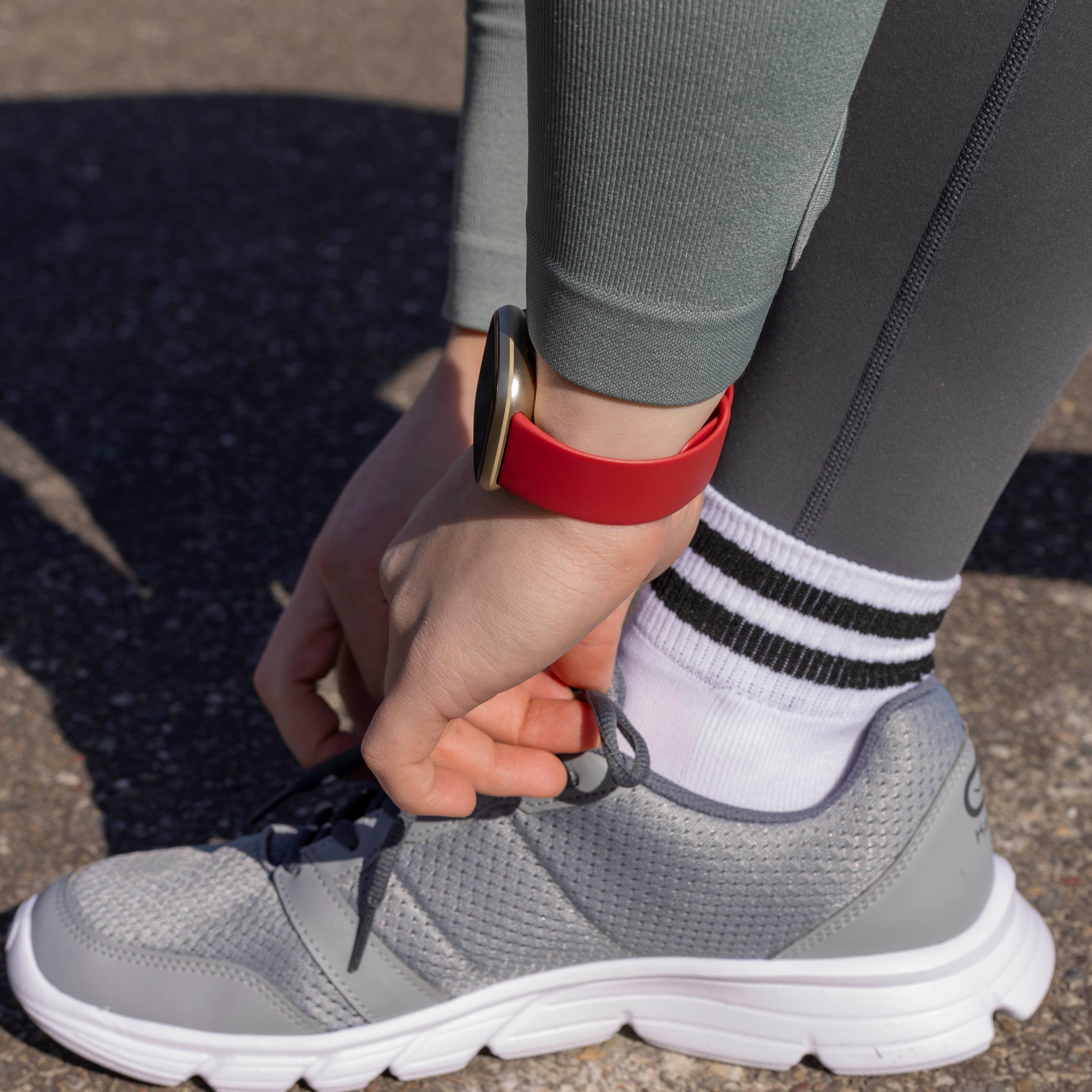 Eine Person bindet ihren Schnürsenkel. Eine rote Uhr befindet sich am Handgelenk. Die Person trägt graue Leggings, ein grünes Hemd und eine weiße Socke.