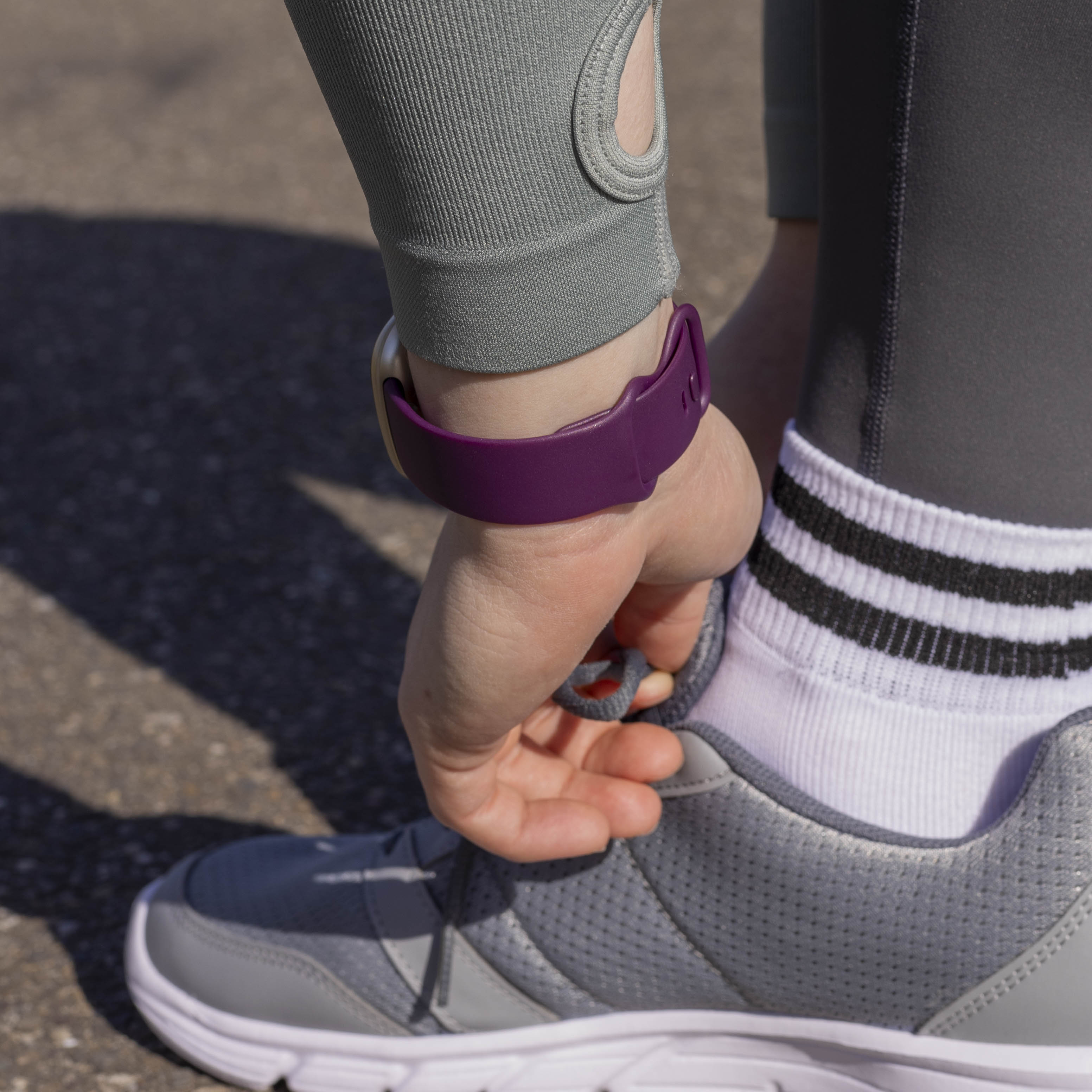 Eine Person bindet einen grauen Laufschuh. Sie trägt graue Leggings, eine lila Uhr und weiße Socken mit schwarzen Streifen.