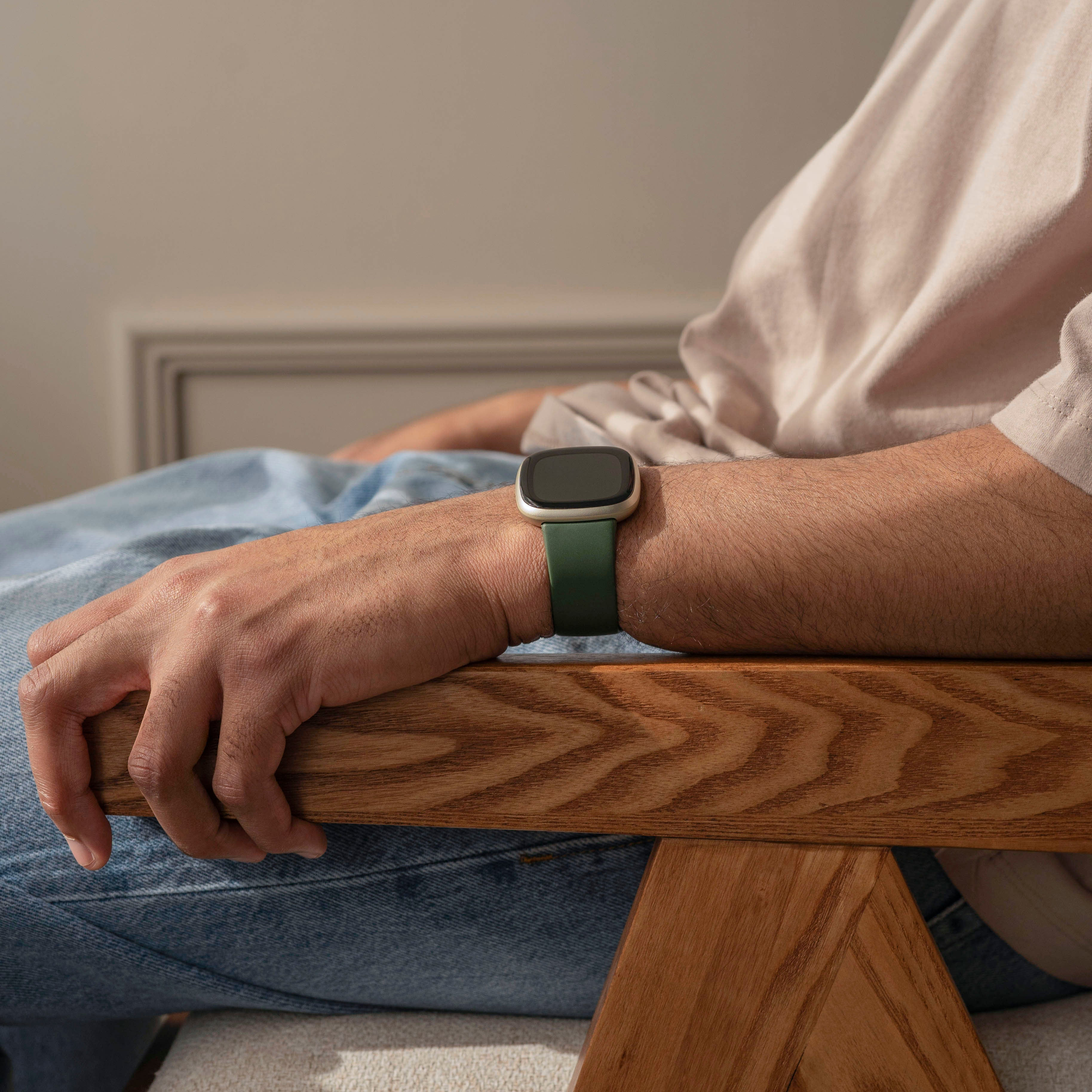 Eine Person trägt eine grüne Smartwatch am Handgelenk und sitzt auf einem Holzstuhl. Helles Hemd und blaue Jeans sind sichtbar.