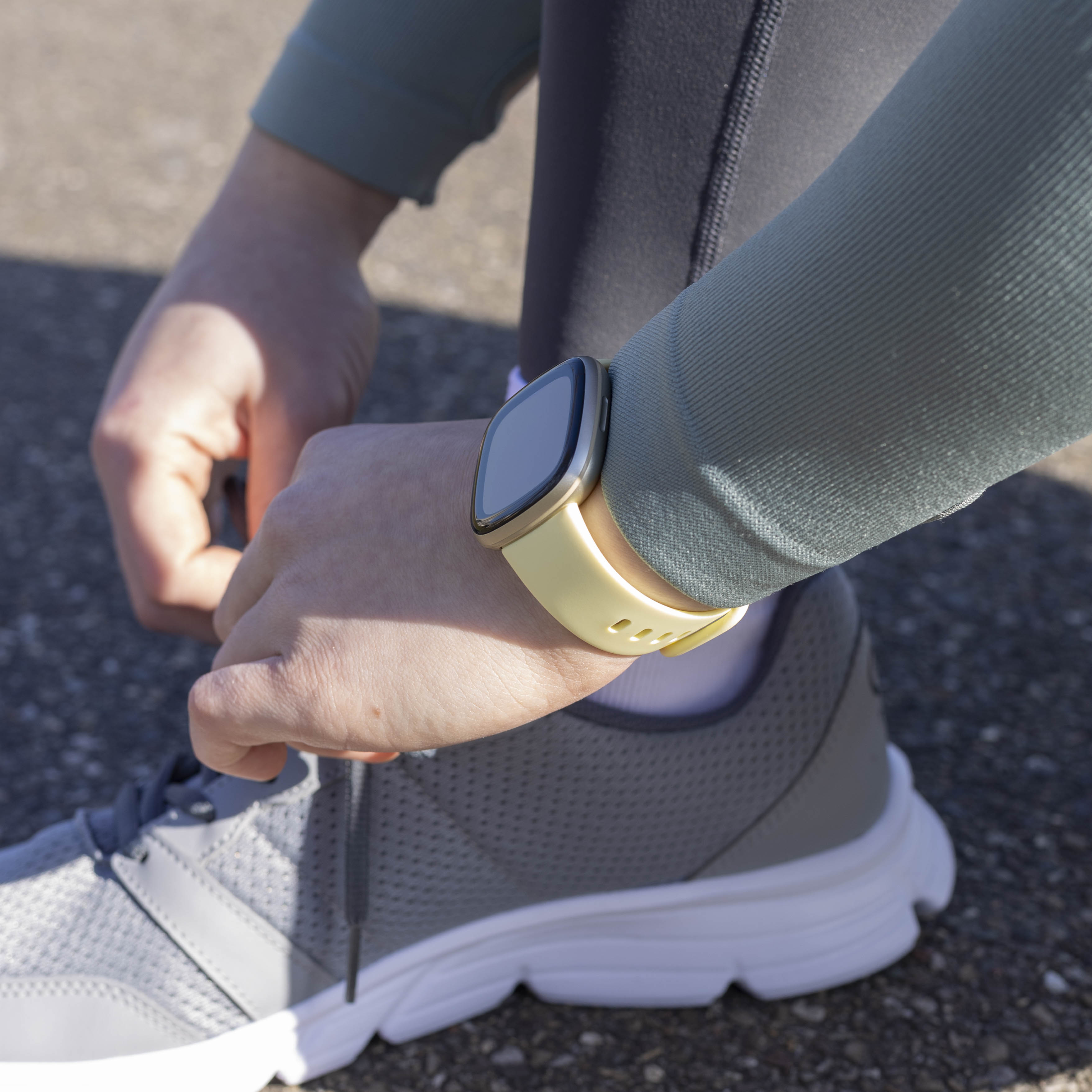 Person schnürt einen grauen Laufschuh. Eine Uhr mit gelbem Armband ist am Handgelenk. Die Szene spielt sich im Freien ab.