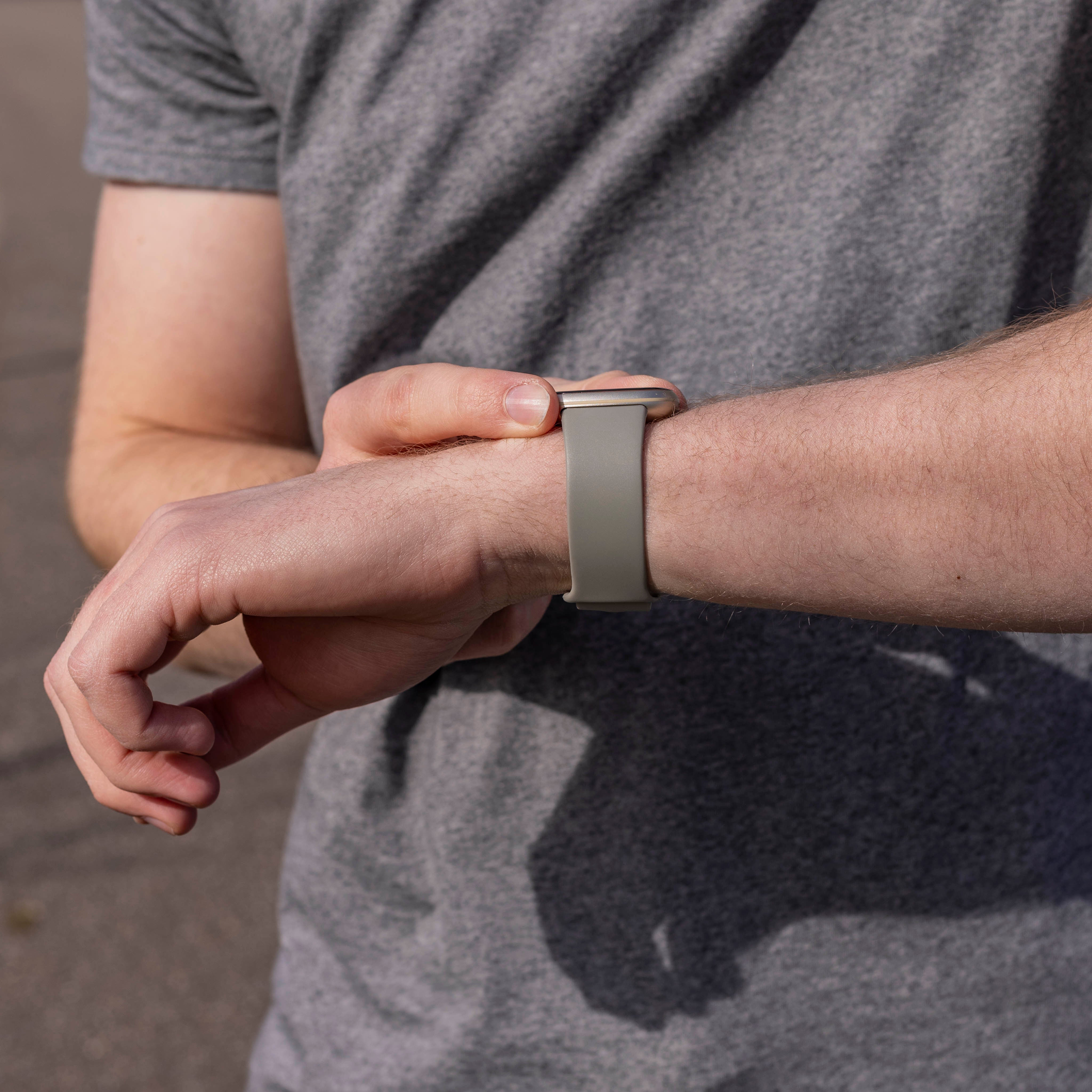 Eine Person passt eine graue Smartwatch an ihrem Handgelenk an. Die Person trägt ein graues Hemd. Der Hintergrund ist verschwommen.