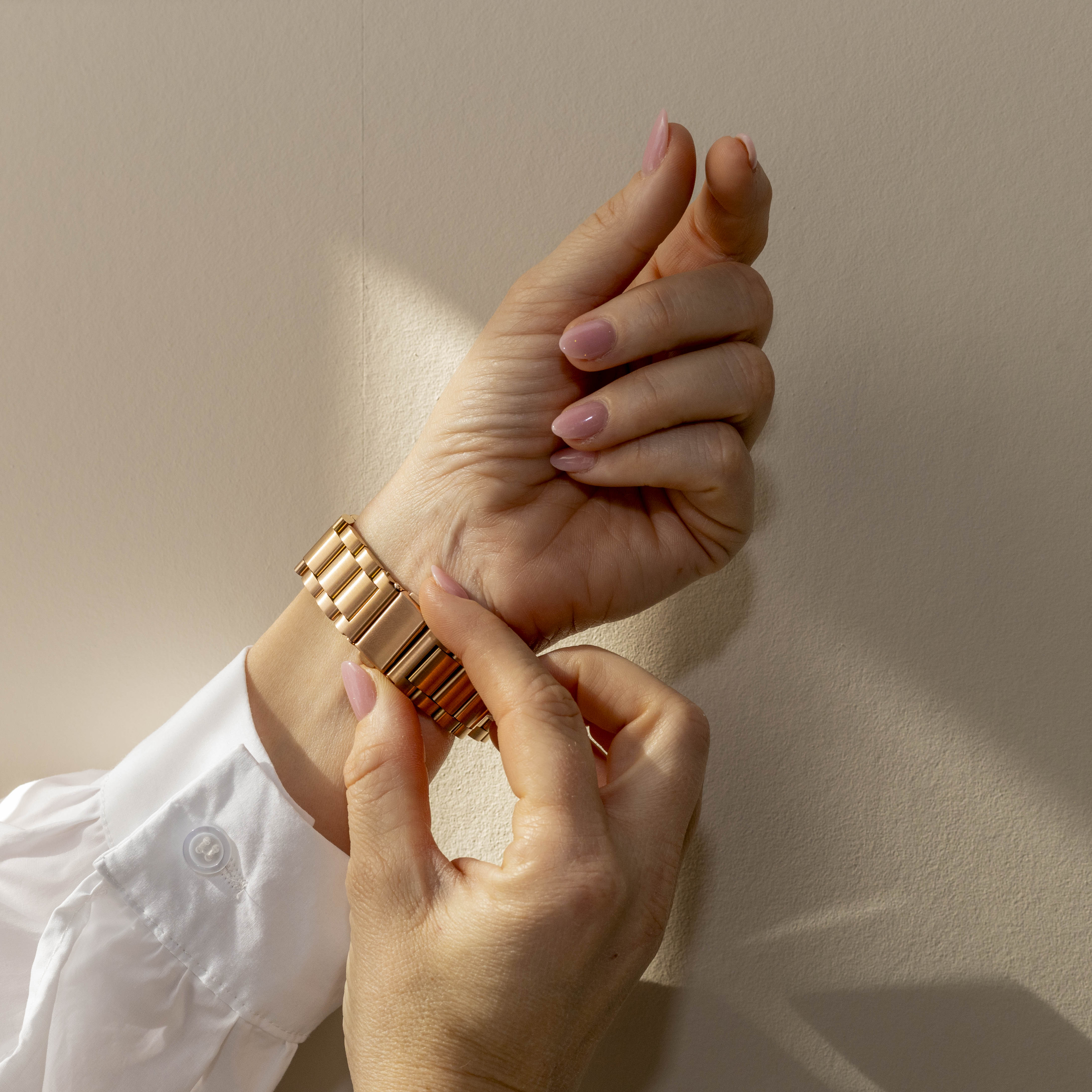 Hände legen eine goldene Uhr an. Die Nägel sind rosa lackiert, das Hemd ist weiß und der Hintergrund ist beige.