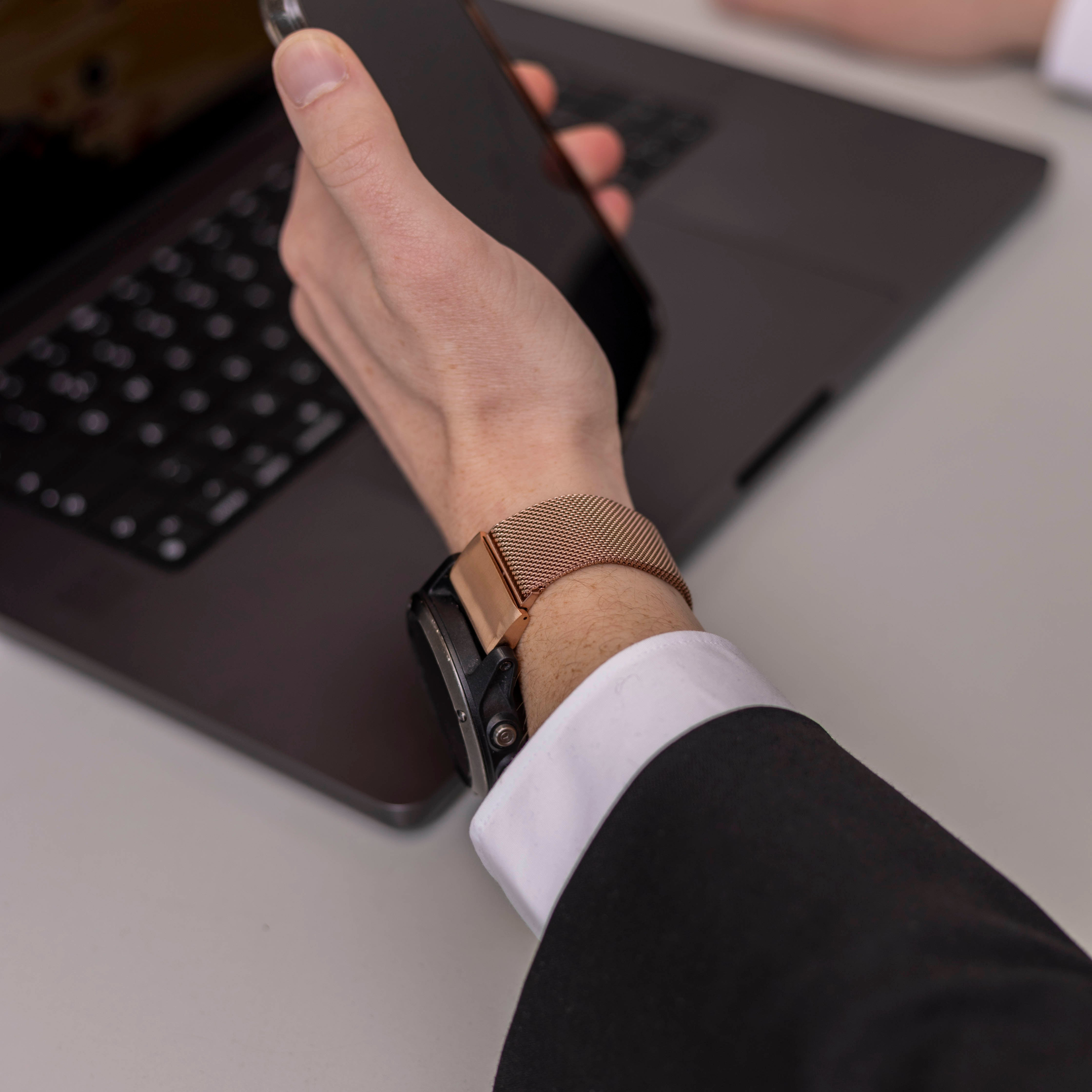 Eine Person, die ein Telefon über einem Laptop hält. Am Handgelenk befindet sich eine roségoldene Uhr. Die Person trägt einen schwarzen Anzug.