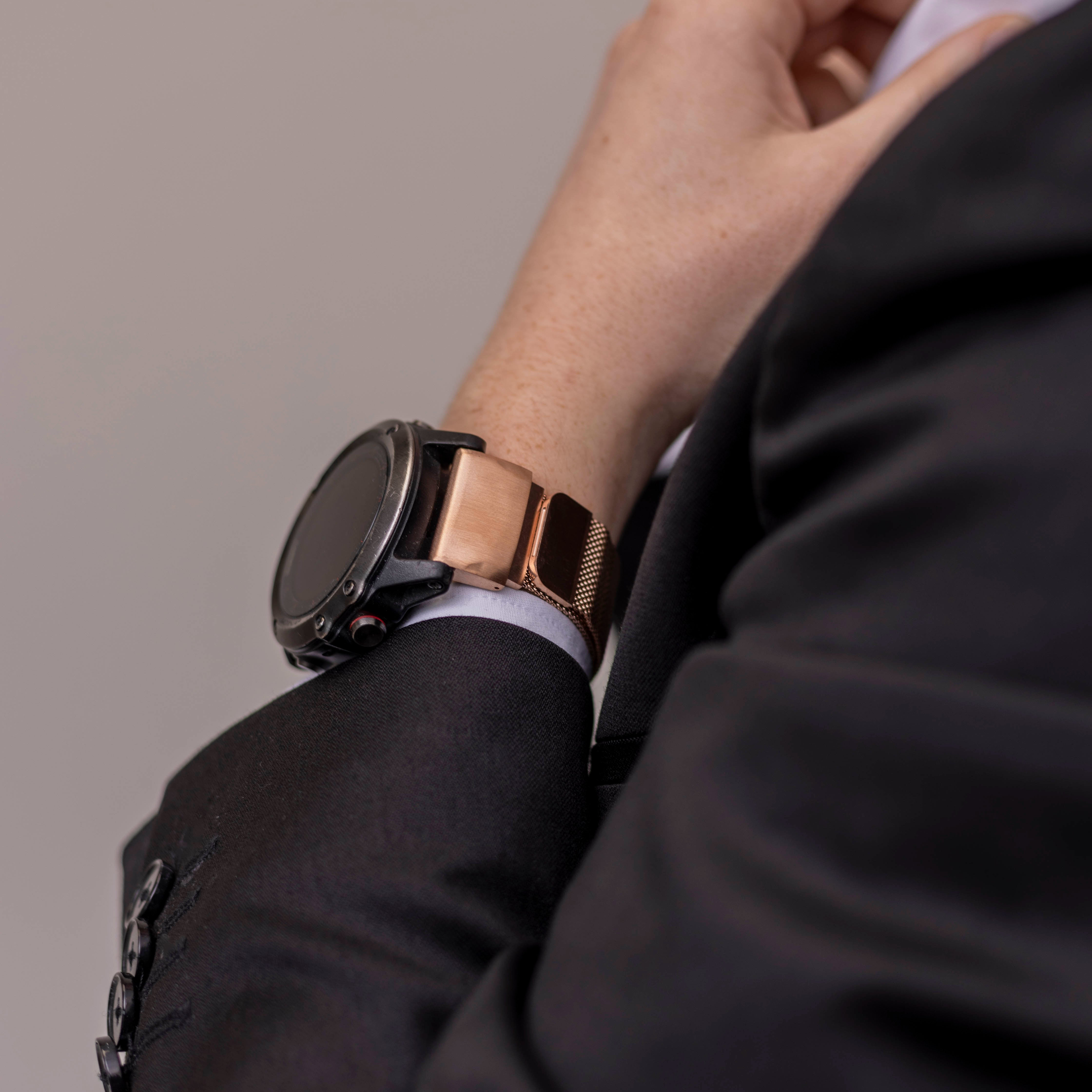Eine Person in einem schwarzen Anzug trägt eine schwarze und roségoldene Smartwatch. Der Hintergrund ist hellgrau. Der Arm der Person ist sichtbar.