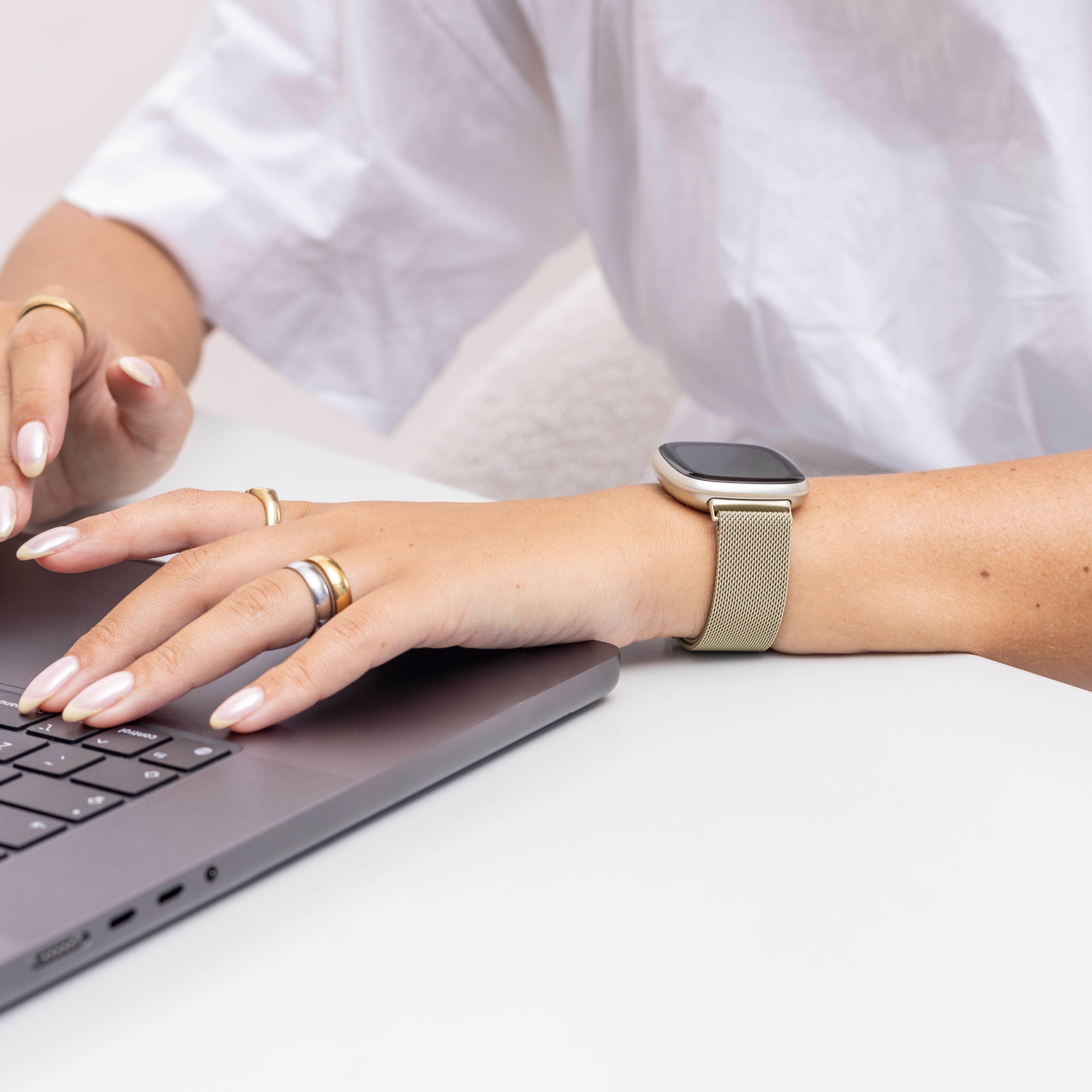 Hände einer Person auf einer Laptop-Tastatur. Sie tragen Ringe und eine Smartwatch. Weißes Hemd auf weißem Tisch.