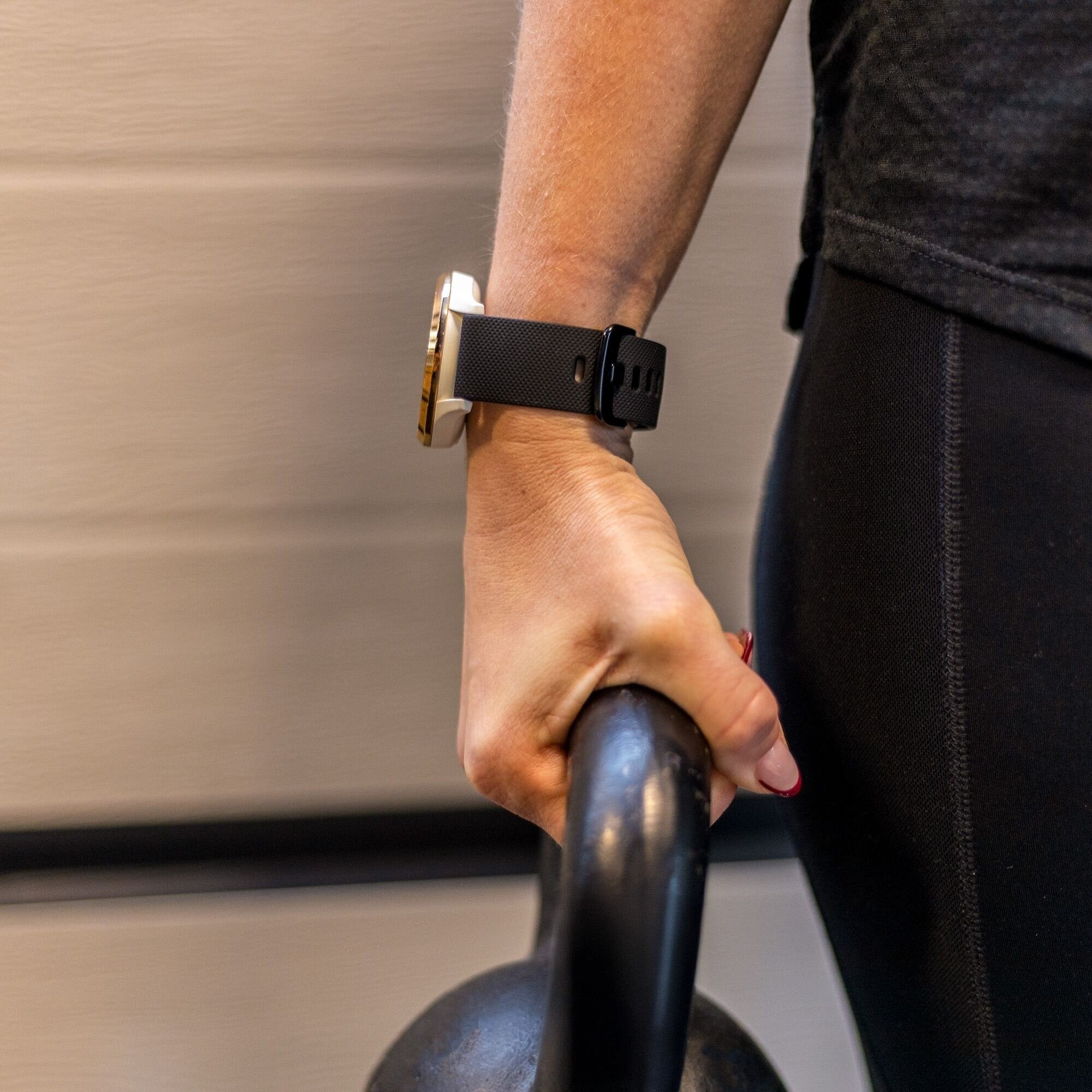 Eine Person hält eine schwarze Kettlebell mit einer Uhr am Handgelenk. Sie trägt schwarze Leggings und ein dunkles Oberteil.