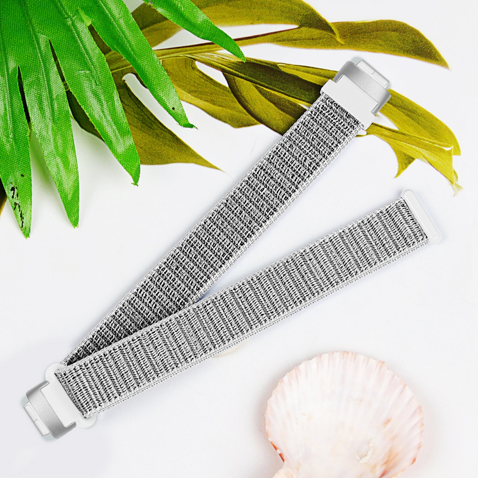Ein Uhrenarmband mit silberner Hardware, grau gewebtem Stoff und einer Muschel. Grüne und gelbe Blätter im Hintergrund.