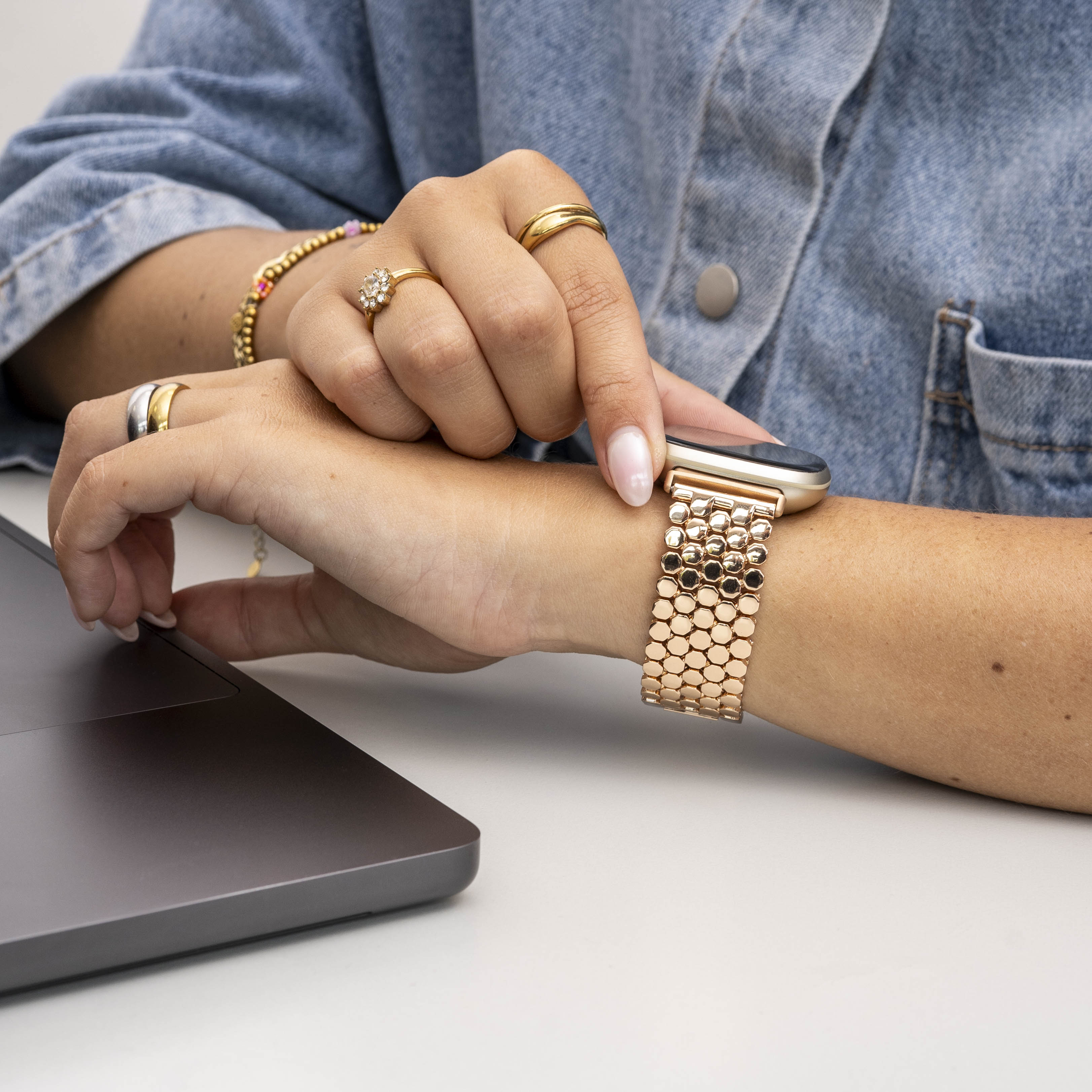 Das Handgelenk einer Person mit einer goldenen Uhr, geschmückt mit Ringen und einem Laptop. Die Person trägt eine Jeansjacke.