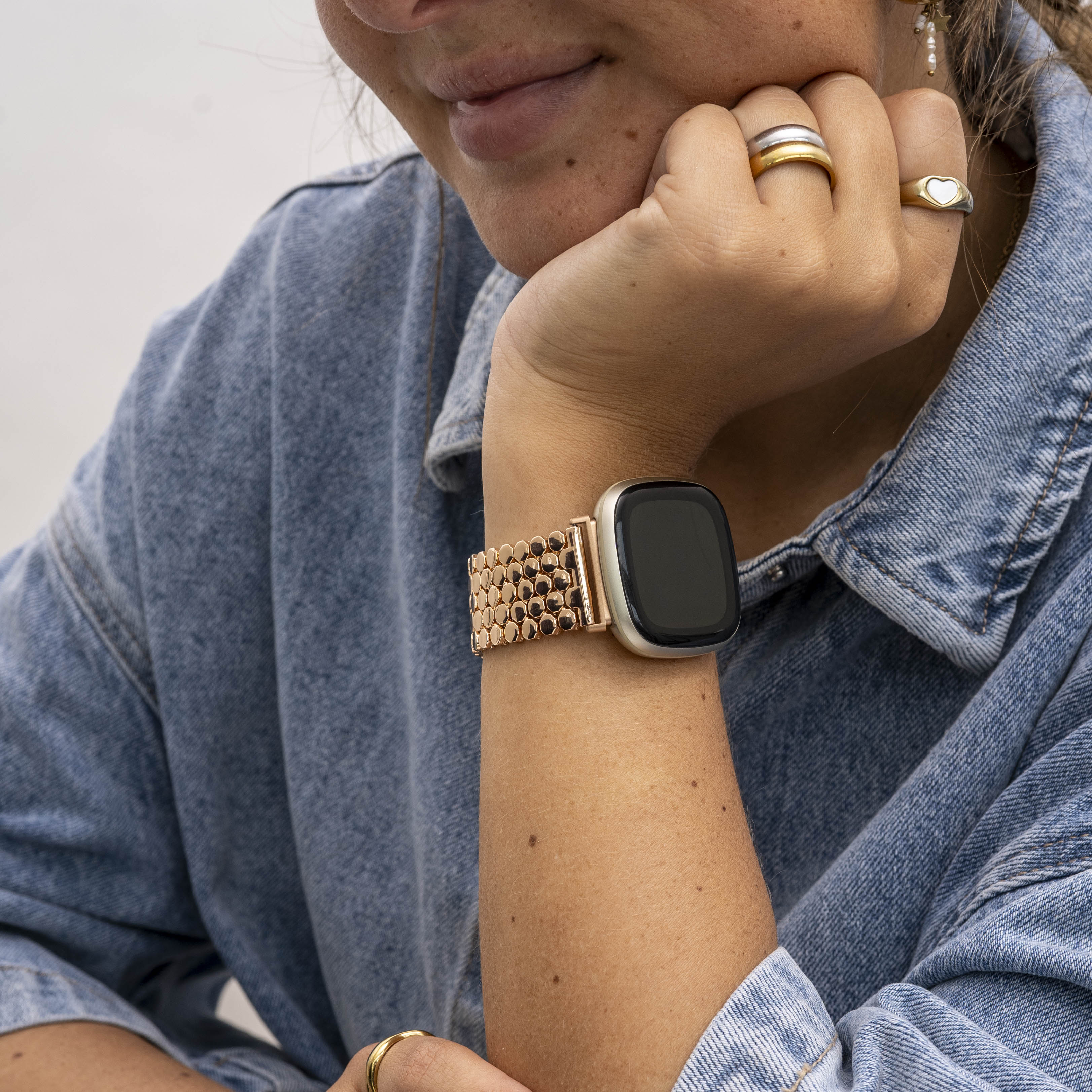Eine Frau mit Ringen und einer goldenen Uhr. Sie trägt eine Jeansjacke und stützt das Kinn auf die Hand.