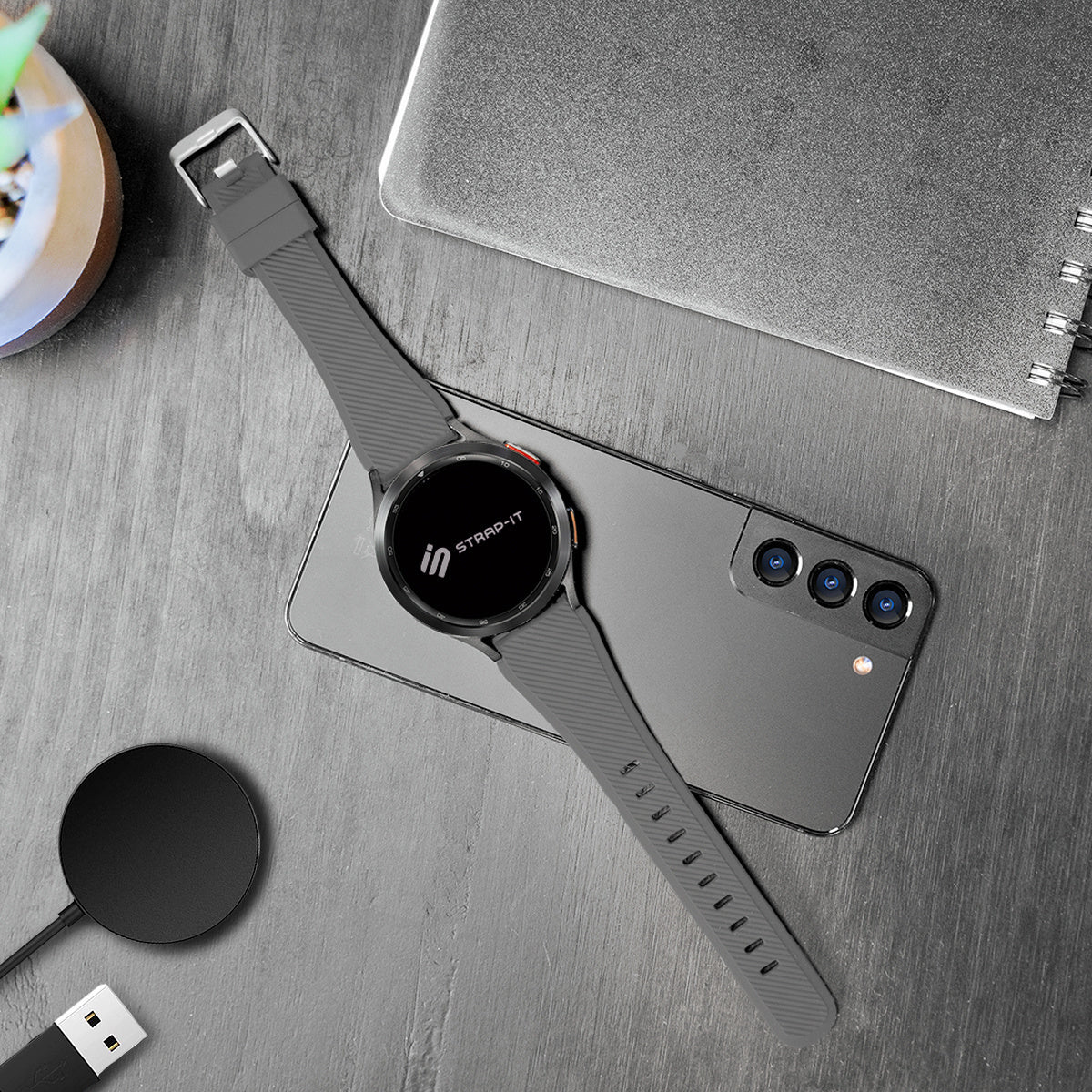 Eine graue Smartwatch auf einem grauen Telefon, ein Ladepad und ein Notizbuch liegen auf einem Holztisch. Die Uhr hat einen schwarzen Bildschirm.