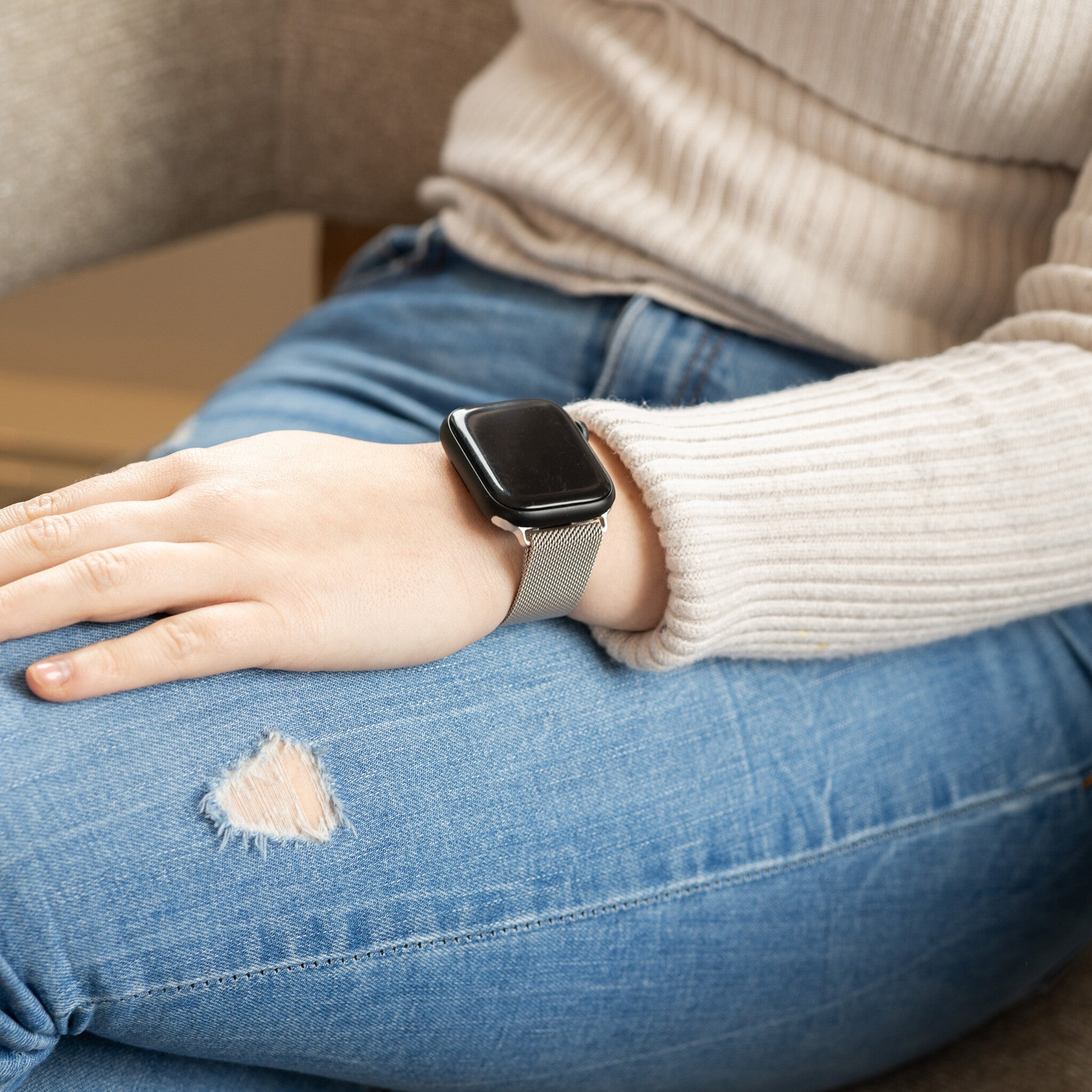 Eine Person mit beigem Pullover und Jeans sitzt. Eine Uhr mit silbernem Armband am Handgelenk.