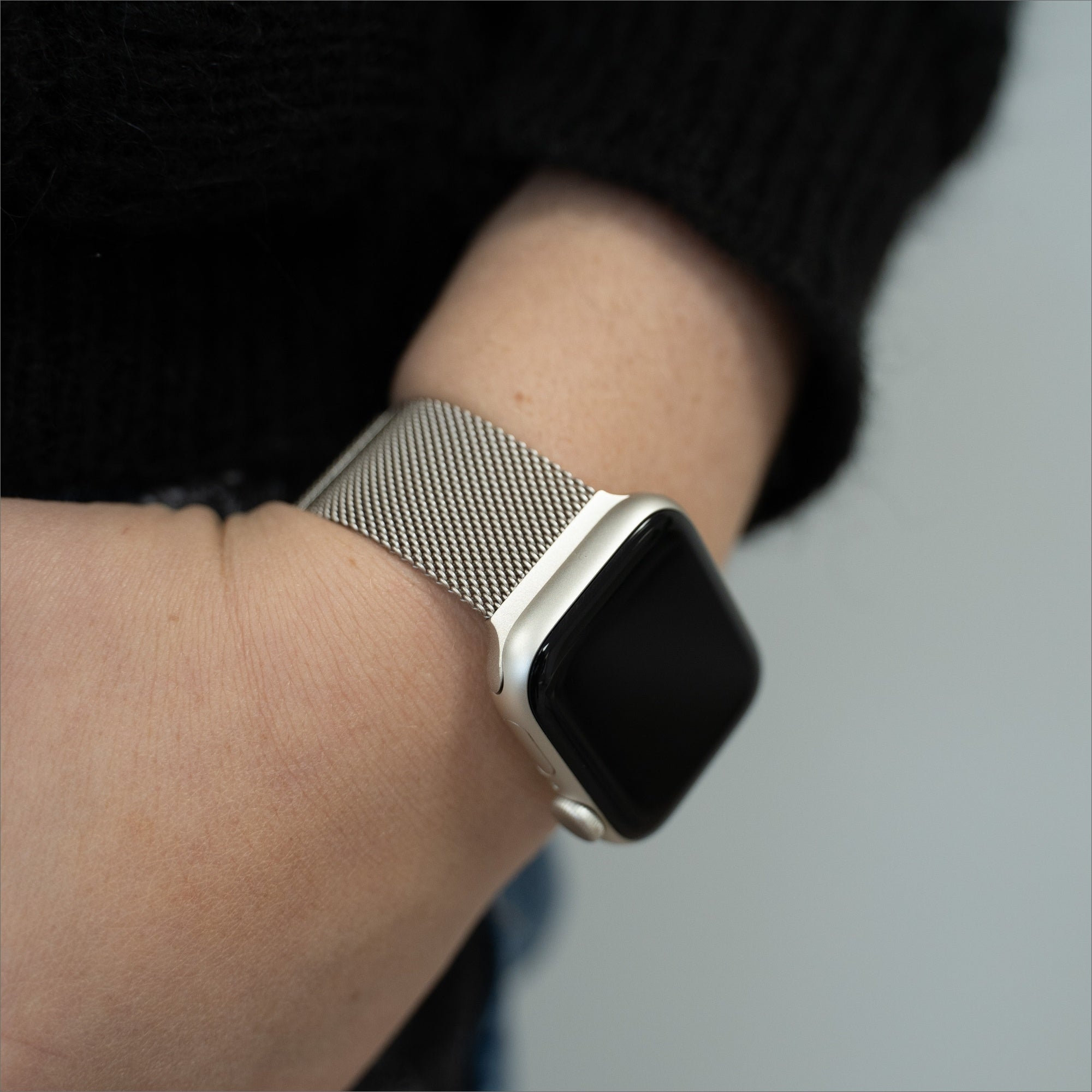 Ein Handgelenk mit einer silbernen Apple Watch, einem Mesh-Armband und einem schwarzen Bildschirm. Der Arm ist in einem schwarzen Pullover. Grauer Hintergrund.