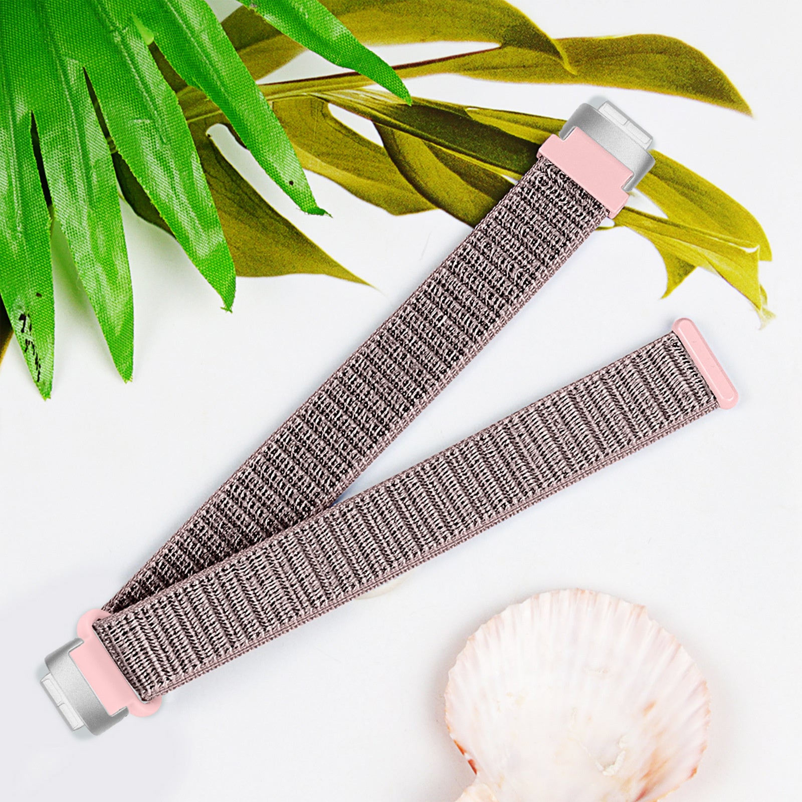 Ein Uhrenarmband mit rosa Akzent wird mit grünen Blättern und einer Muschel auf weißem Hintergrund angezeigt.