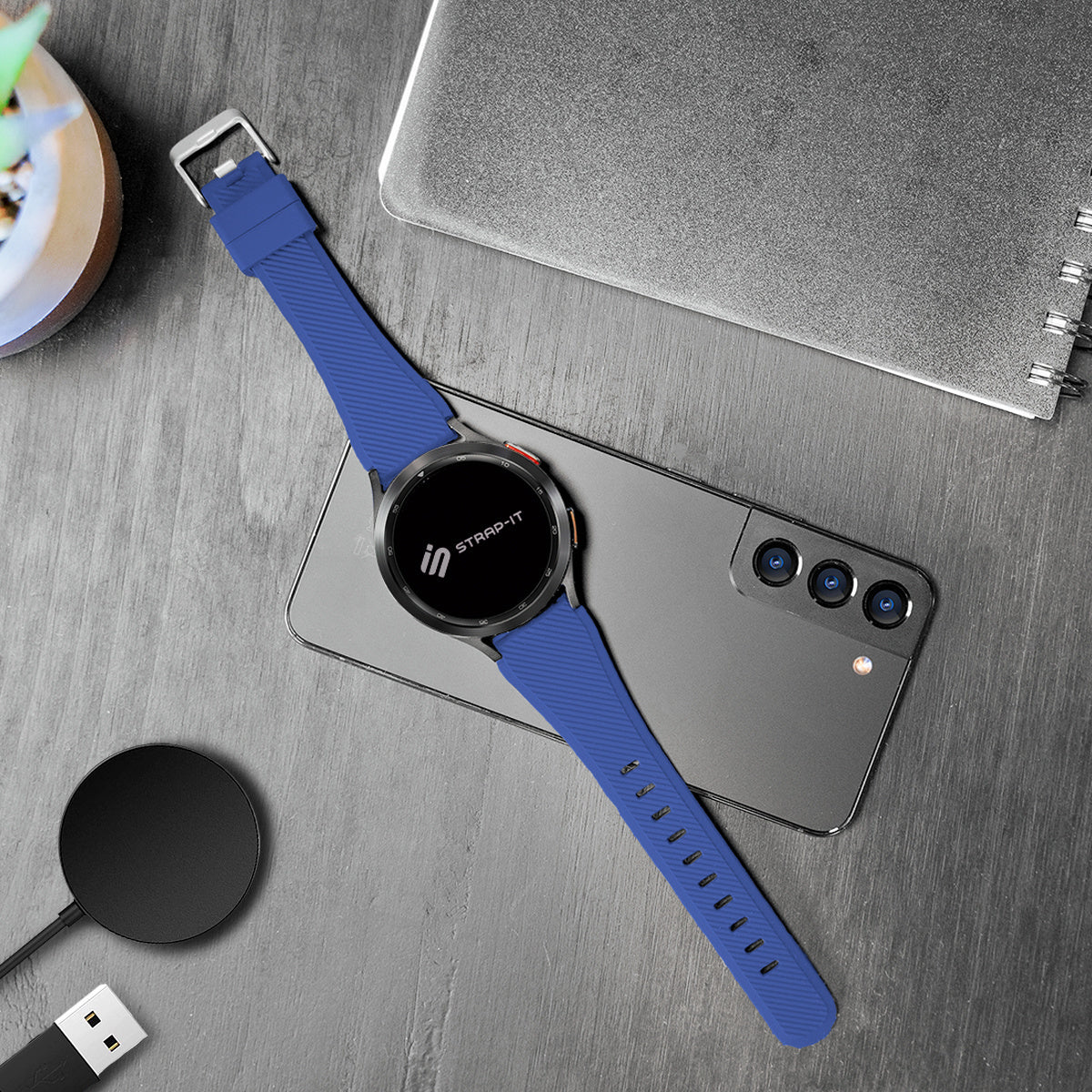 Eine Smartwatch mit blauem Armband auf einem grauen Telefon. Ein Ladegerät und ein Notizbuch sind ebenfalls vorhanden.