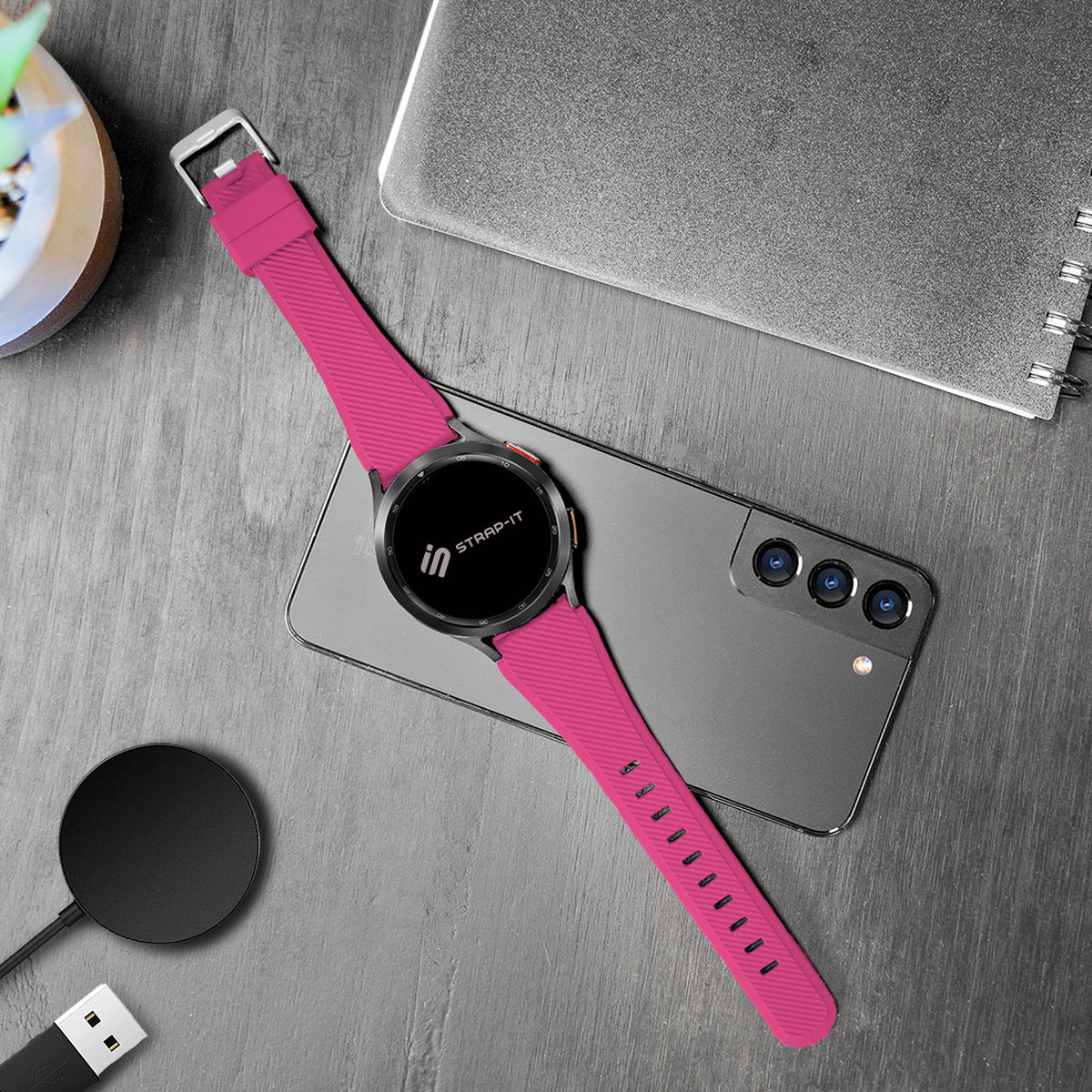Eine pinke Smartwatch liegt auf einem Telefon. Auf einer Holzoberfläche befinden sich auch ein Ladepad und ein Notizbuch.