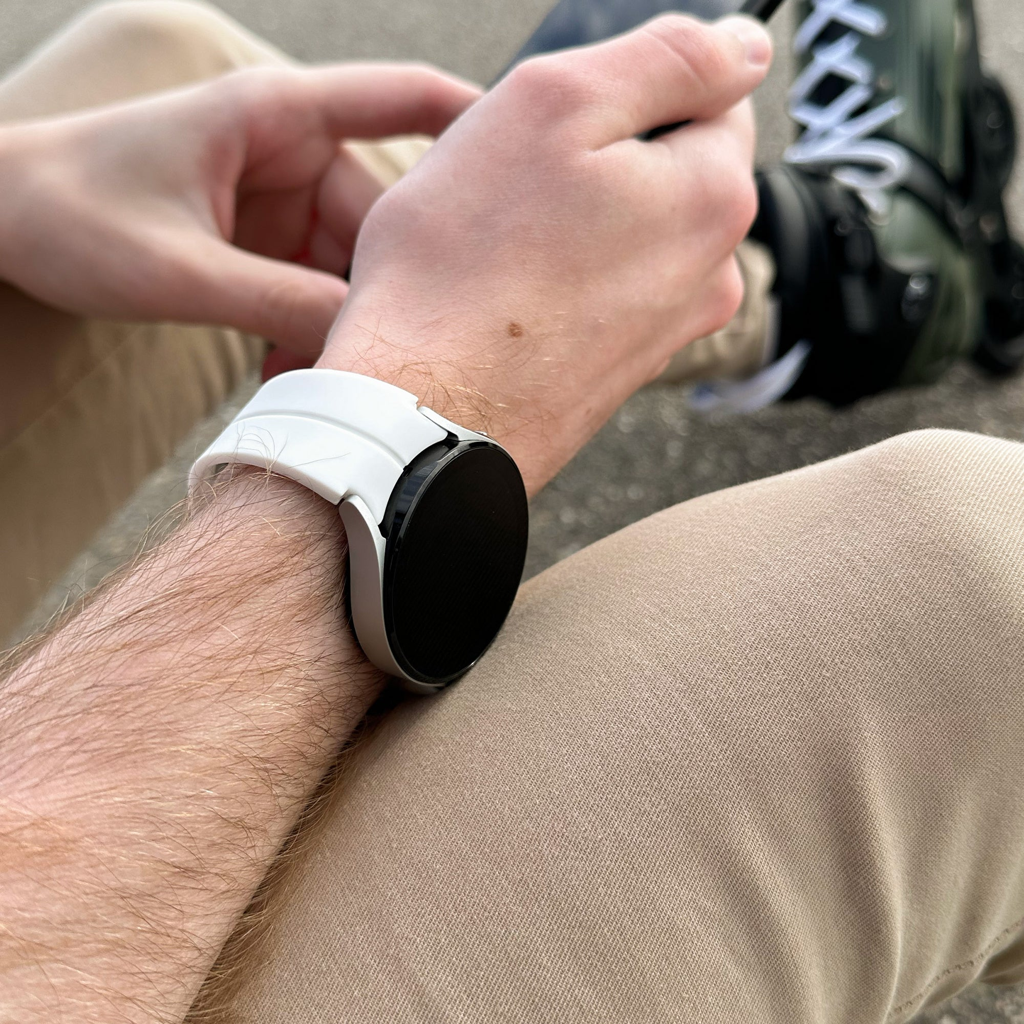 Eine Person trägt eine weiße Smartwatch und sitzt mit beigefarbener Hose da. Im Hintergrund sind Rollschuhe. Die Hand der Person ist sichtbar.