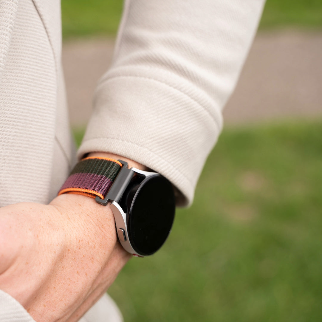 Handgelenk einer Person mit einer Smartwatch. Die Uhr hat einen schwarzen Bildschirm und ein bunt gestreiftes Band. Die Person trägt einen beigen Mantel.