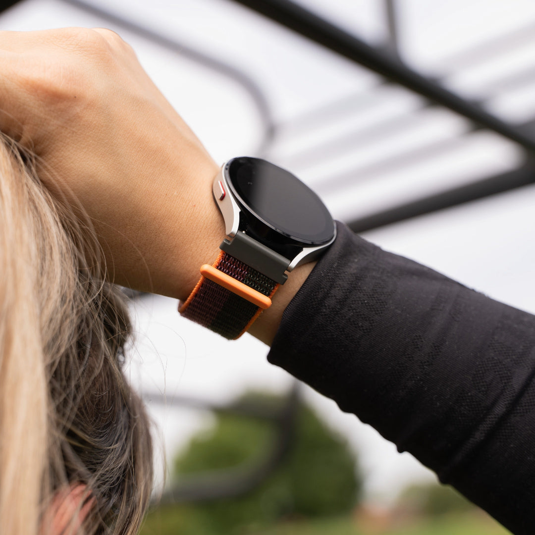 Handgelenk mit Smartwatch. Die Uhr hat einen schwarzen Bildschirm und ein orange-schwarzes Armband. Die Person trägt ein schwarzes Hemd.