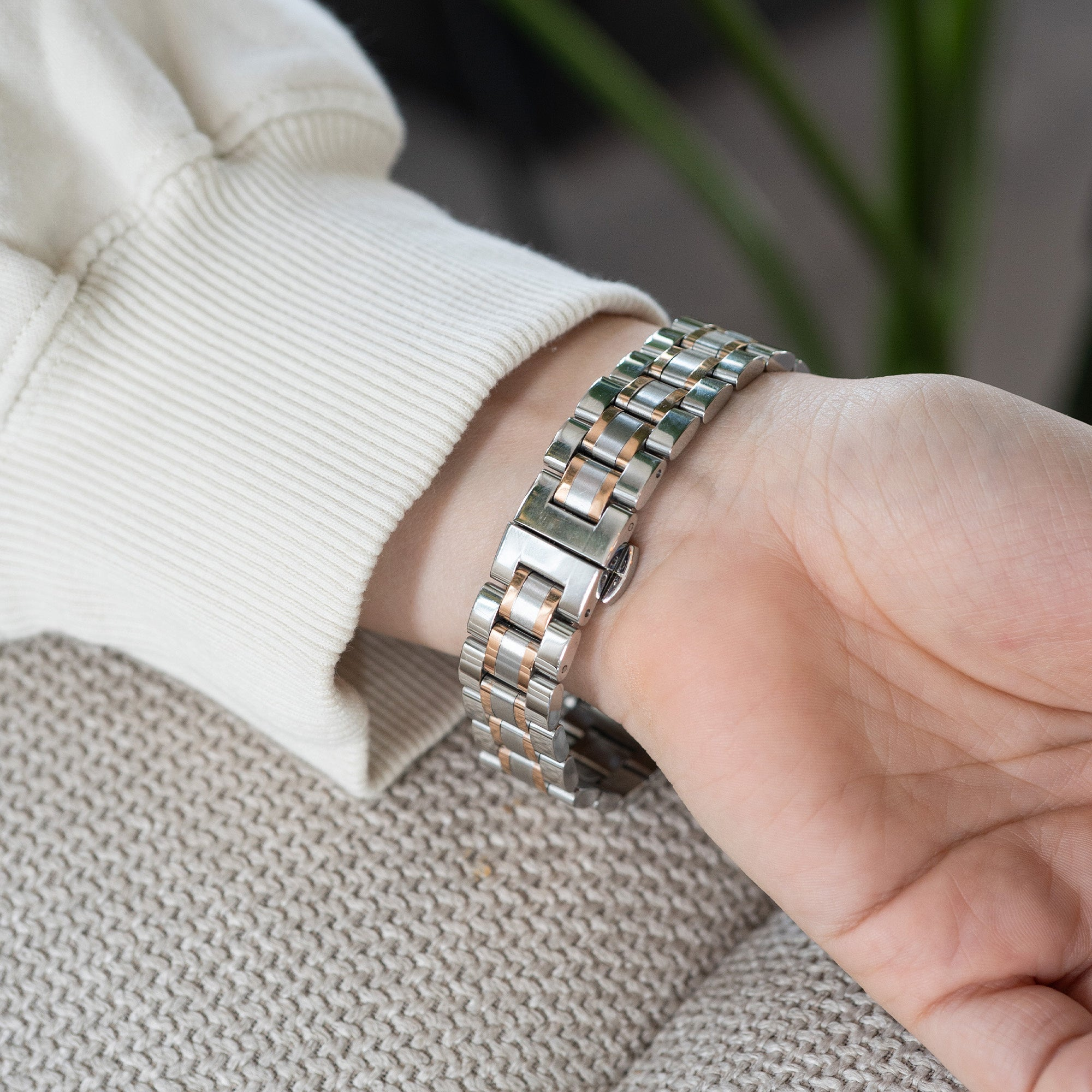 Ein Handgelenk mit einer silber-goldenen Uhr. Der Arm ruht auf einer beige gewebten Oberfläche. Ein weißer Pullover ist sichtbar.