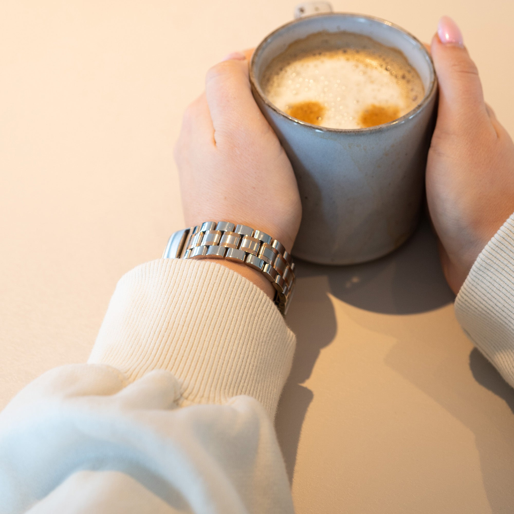 Hände halten eine Tasse Kaffee mit Schaum. Am Handgelenk befindet sich eine Uhr. Beiger Hintergrund.