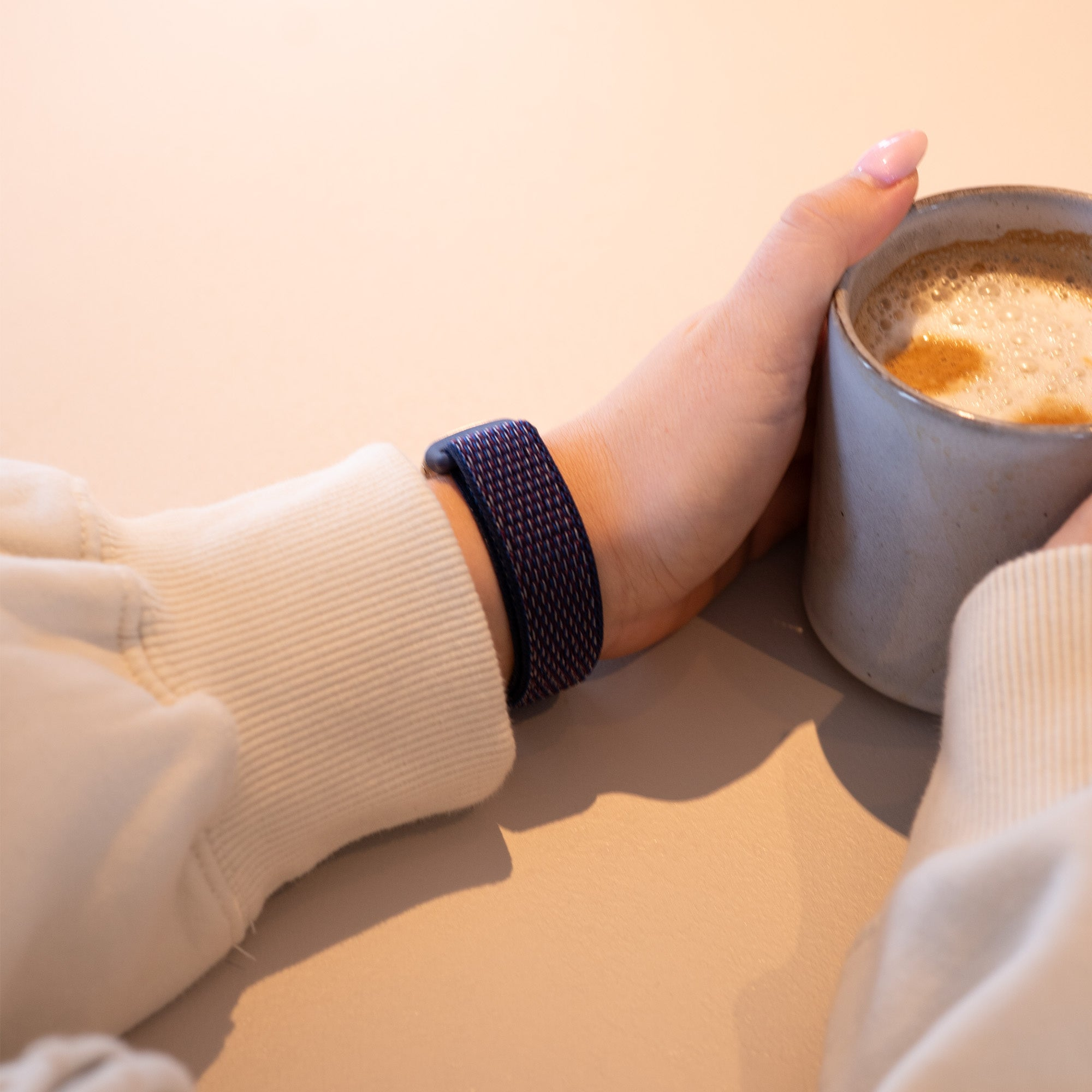 Eine Person hält eine Kaffeetasse. An ihrem Handgelenk befindet sich ein dunkelblaues Band, und sie trägt einen weißen Pullover. Der Hintergrund ist hell.