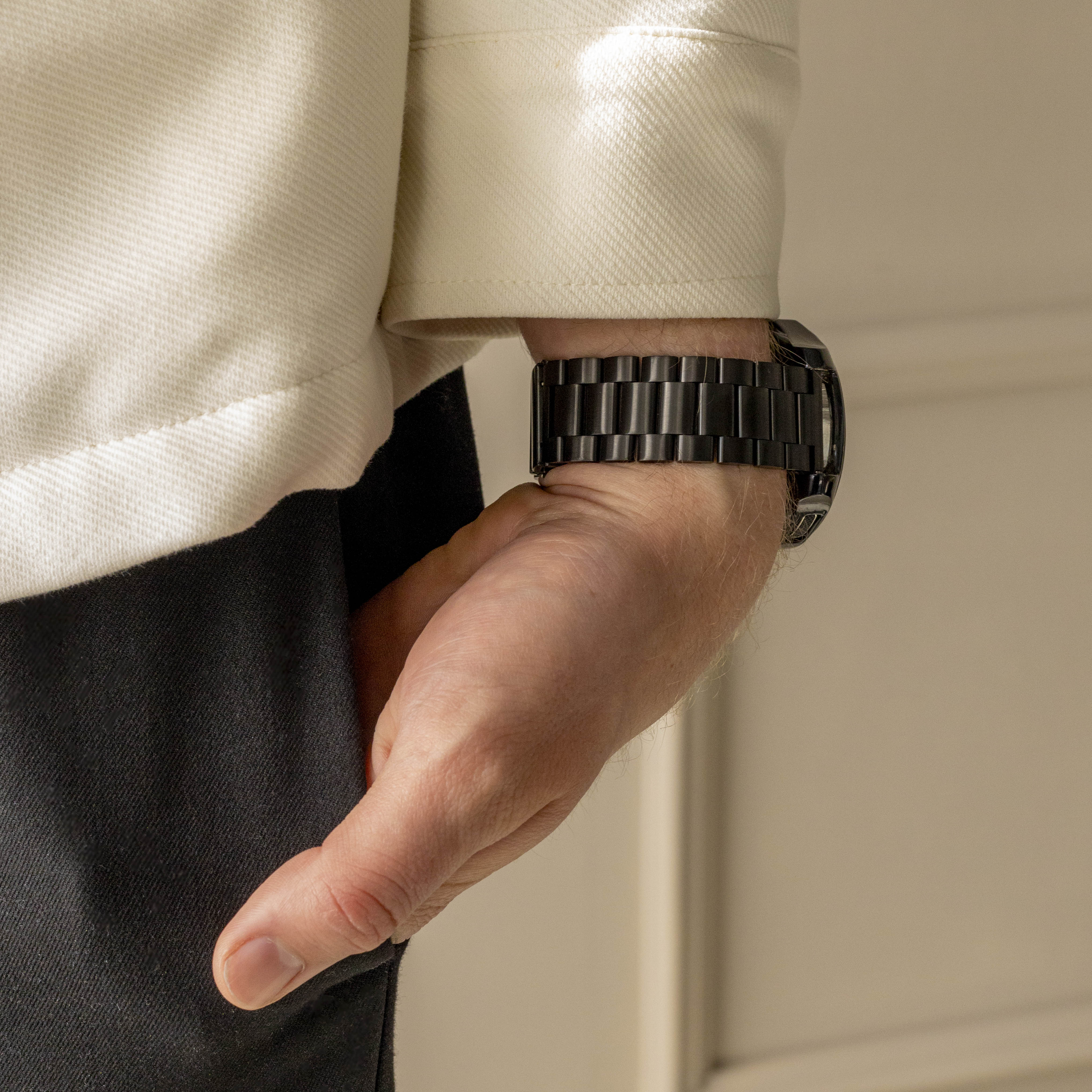 Ein Arm mit schwarzer Uhr vor cremefarbenem Hemd und schwarzer Hose.