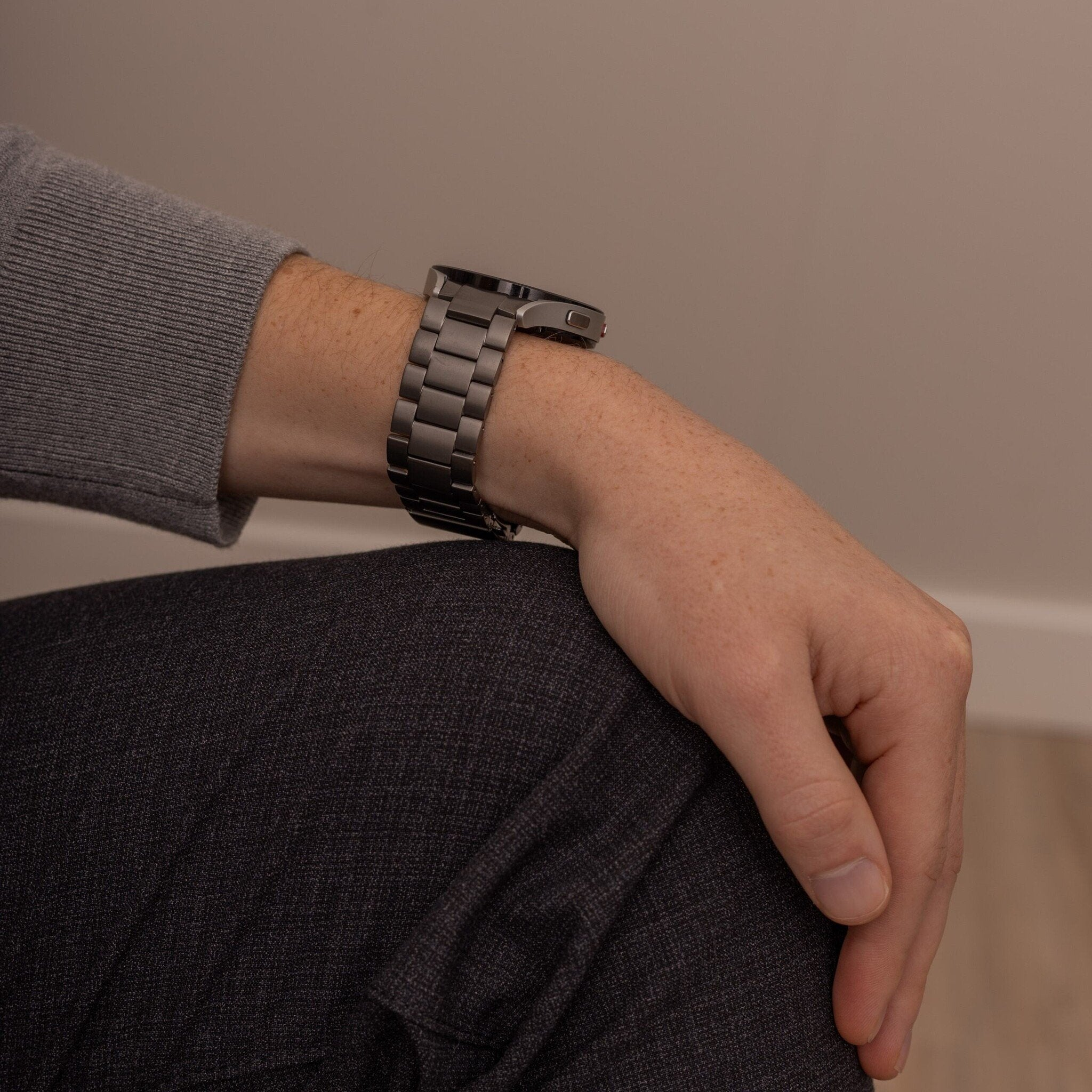 Ein Arm mit einer dunklen Metalluhr. Grauer Pullover, dunkle Hose und eine helle Wand im Hintergrund. Sommersprossen auf dem Arm.