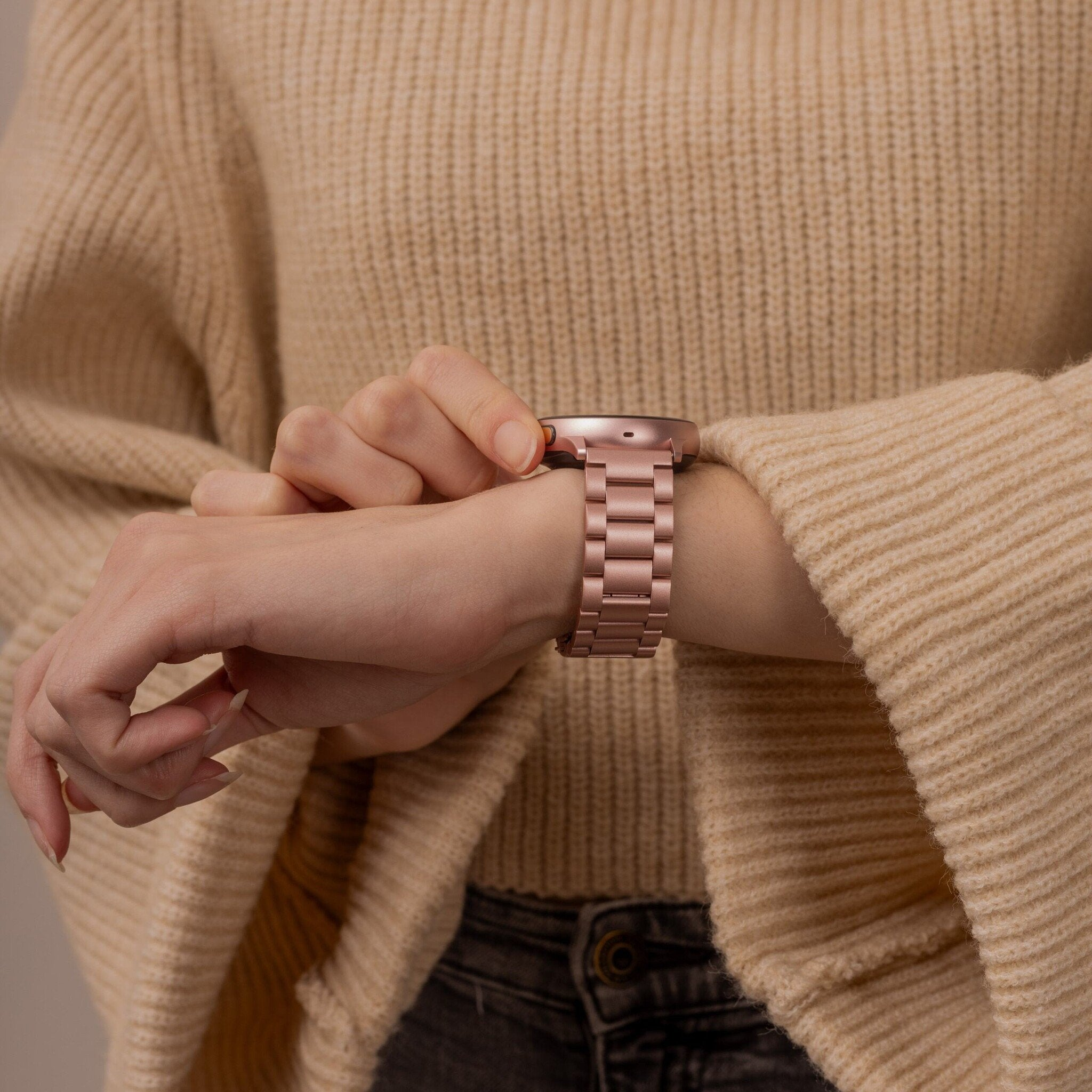 Eine Person, die eine roségoldene Uhr trägt. Die Person trägt einen beigefarbenen Pullover und dunkle Jeans.