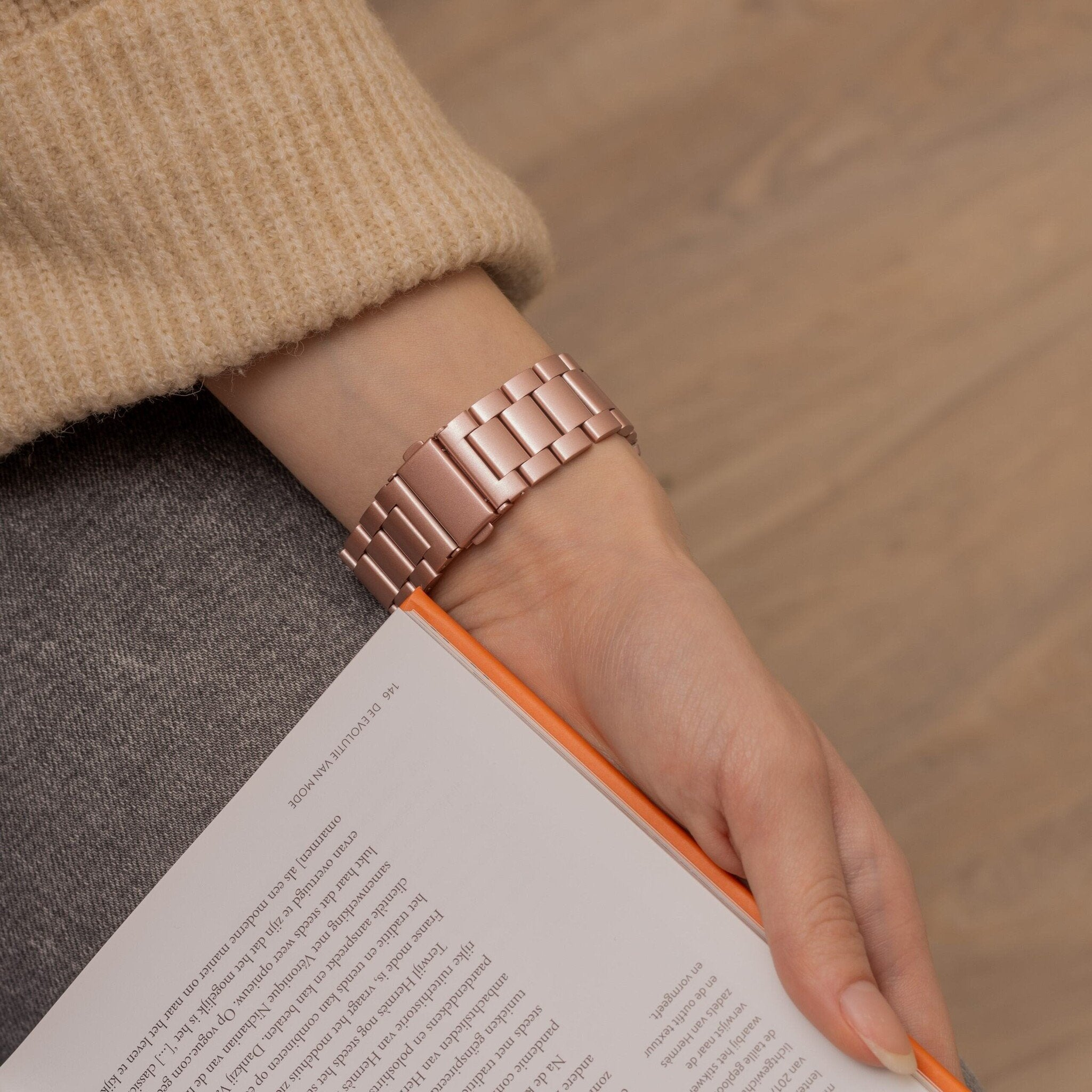 Ein Arm einer Person mit einer roségoldenen Uhr und einem Buch. Die Person trägt einen beigefarbenen Pullover und graue Hosen.