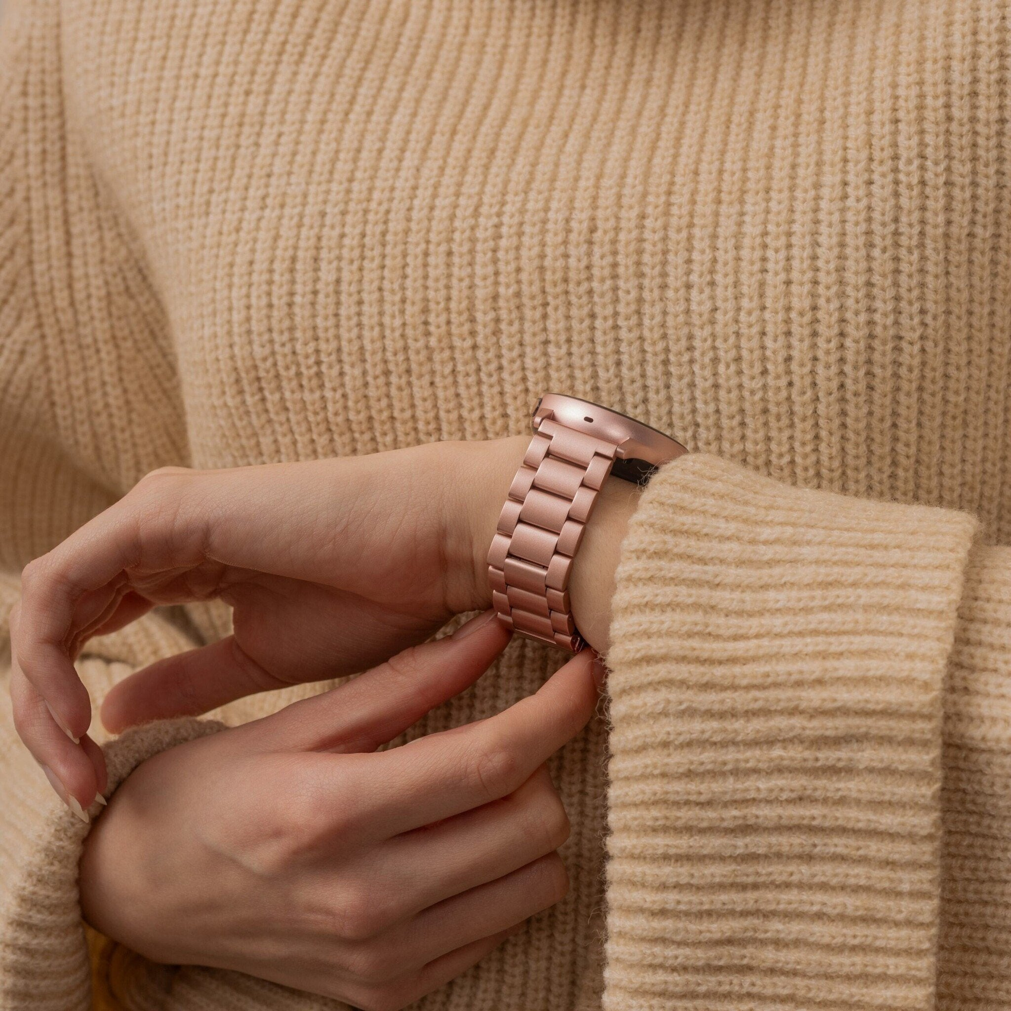 Jemand justiert eine roségoldene Uhr an ihrem Handgelenk und trägt einen beigefarbenen Pullover. Der Hintergrund ist der Stoff des Pullovers.