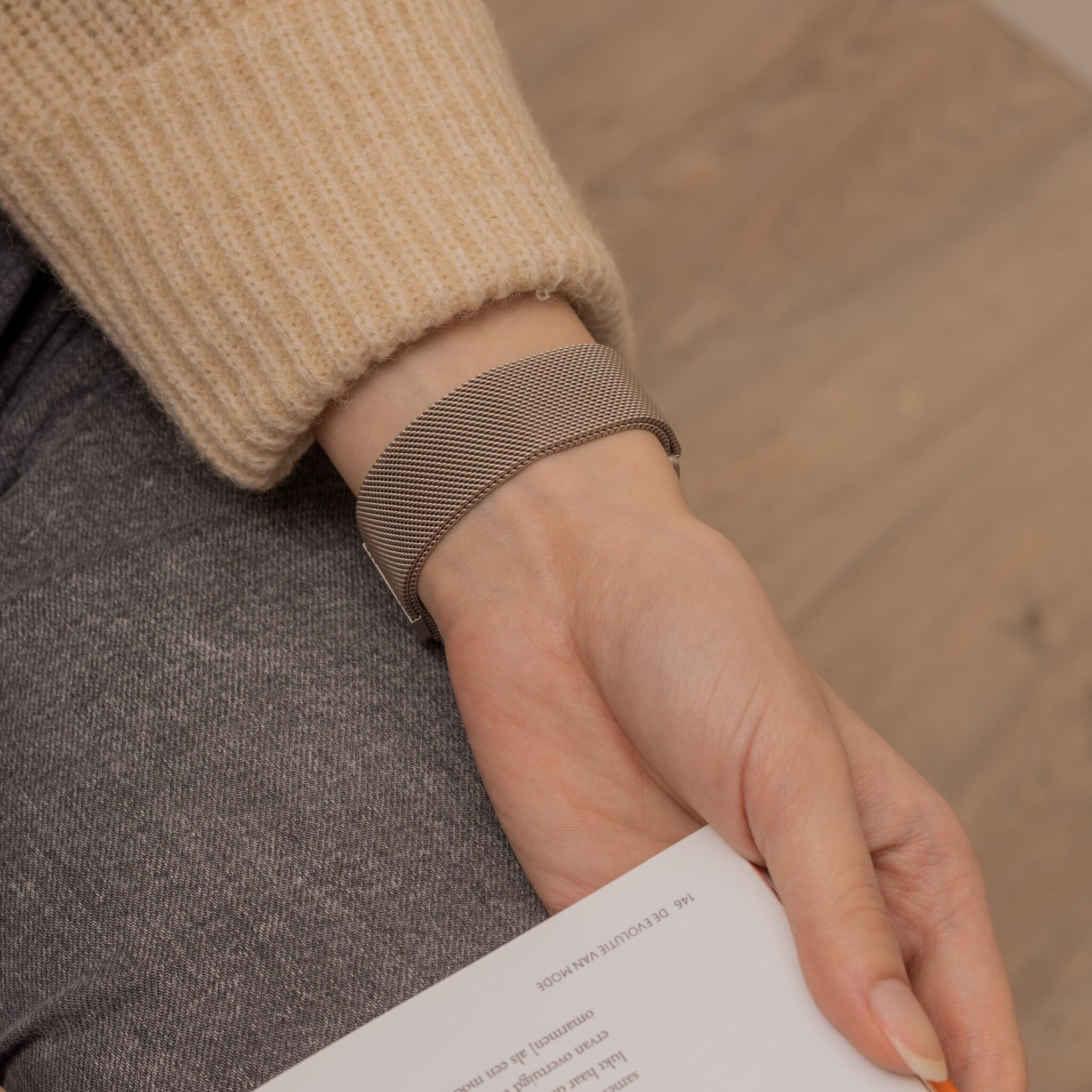 Die Hand einer Person hält ein Papier, trägt ein Metallarmband und einen beigen Pullover. Graue Hose ist sichtbar.