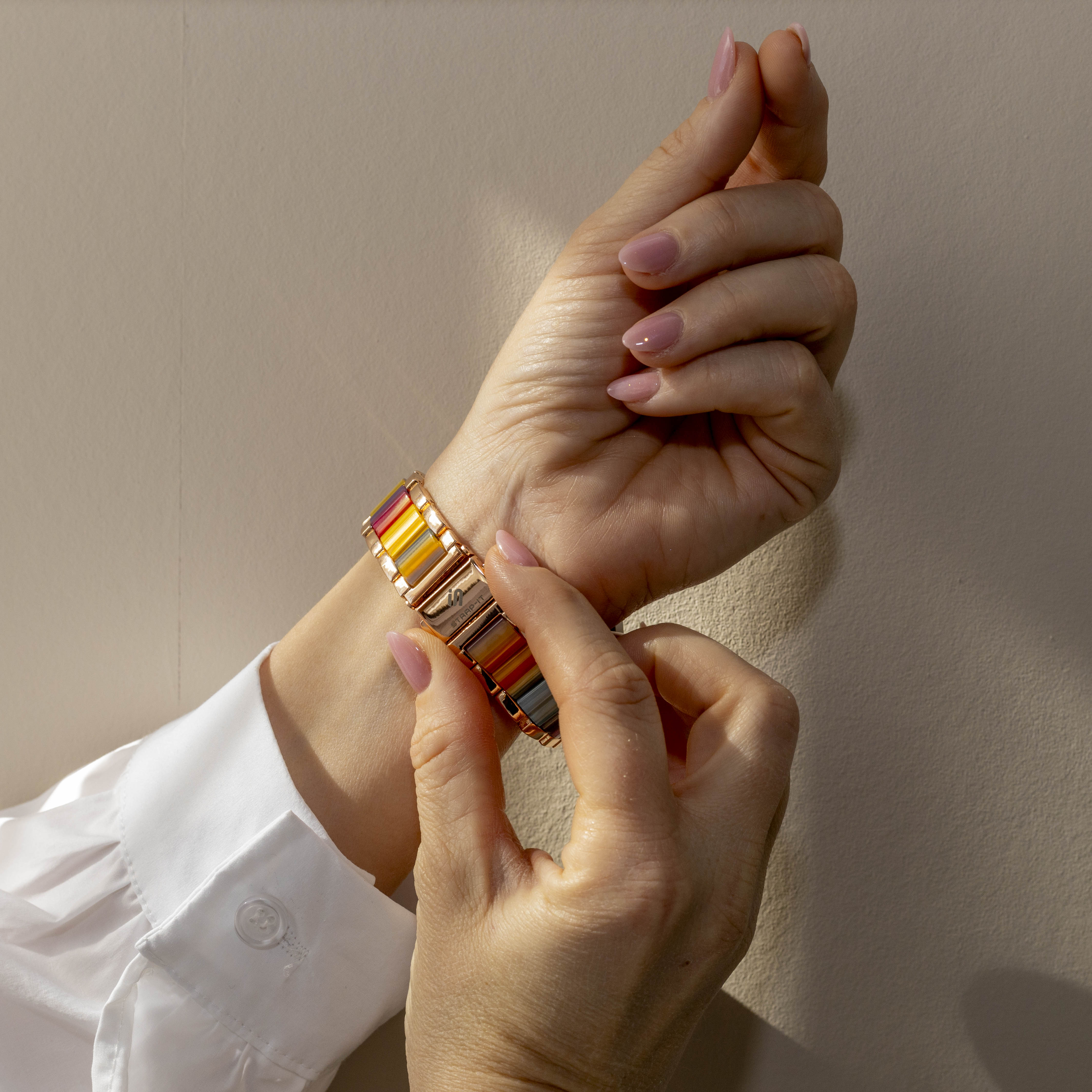 Hände einer Frau legen ein buntes Armband an. Der Hintergrund ist eine beige Wand, sie trägt ein weißes Hemd.