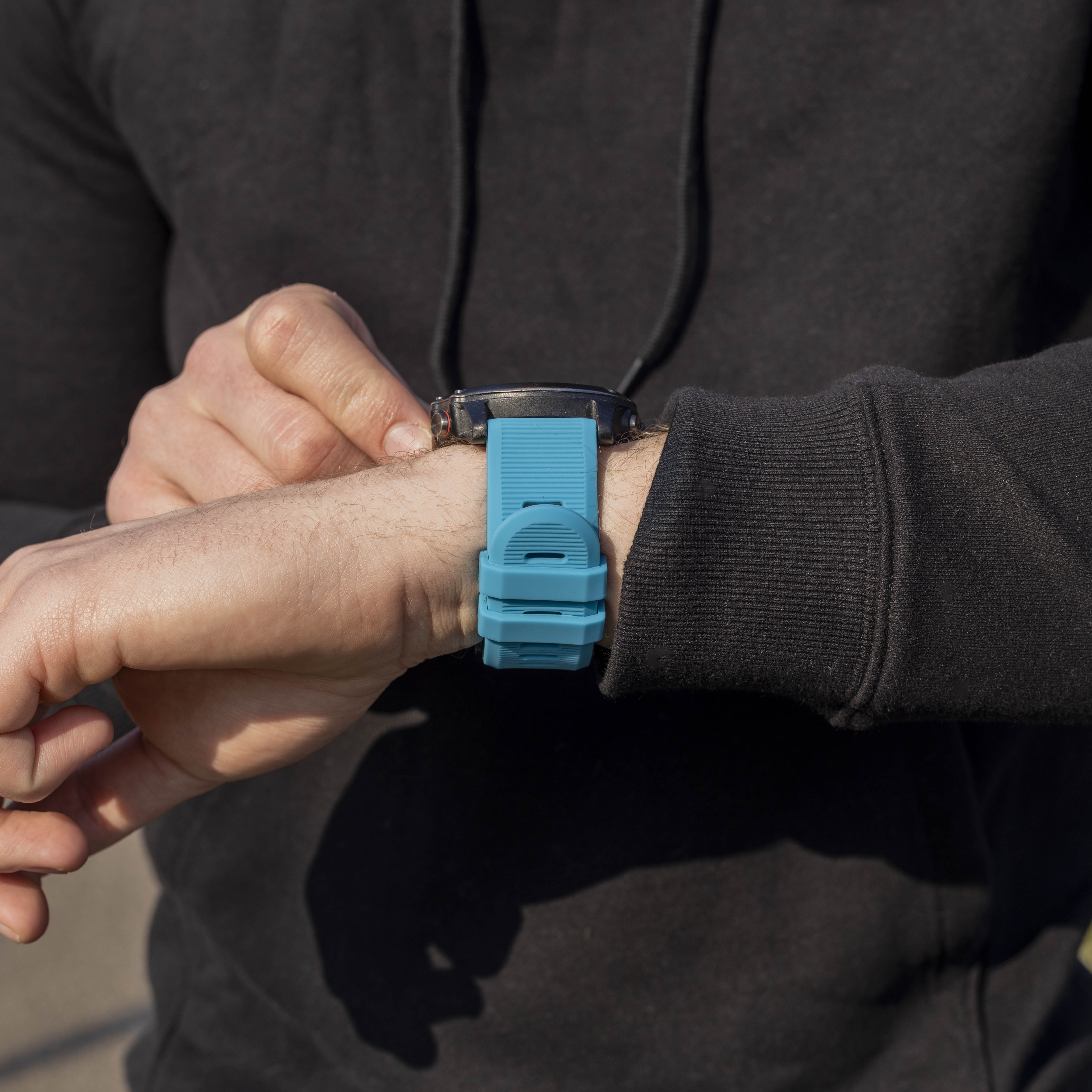 Das Handgelenk einer Person mit einem blauen Uhrenarmband. Die Person trägt einen schwarzen Kapuzenpullover. Die Uhr ist schwarz.