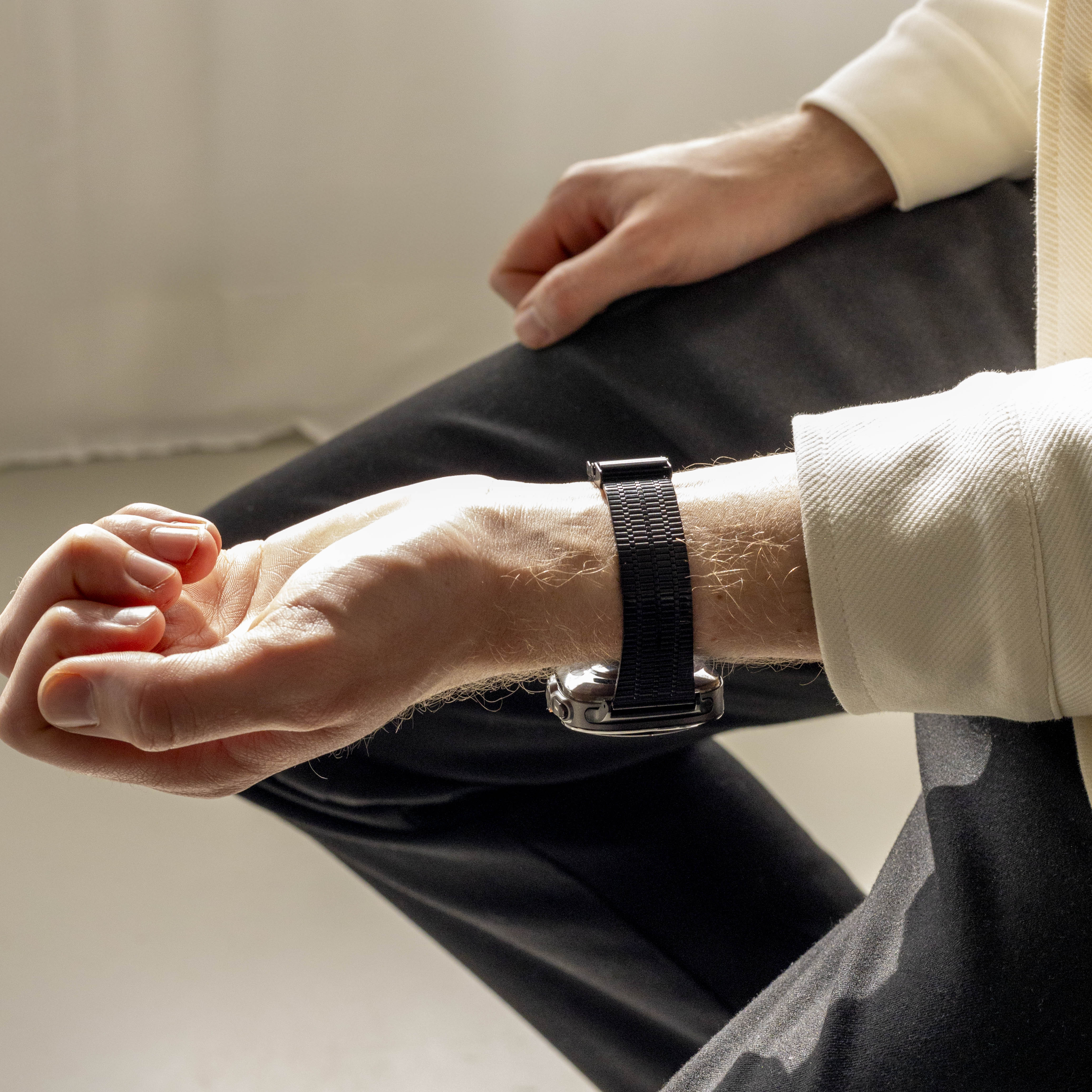 Arm einer Person mit einer Uhr. Die Person trägt eine cremefarbene Jacke und eine schwarze Hose. Die Uhr hat ein schwarzes Armband.