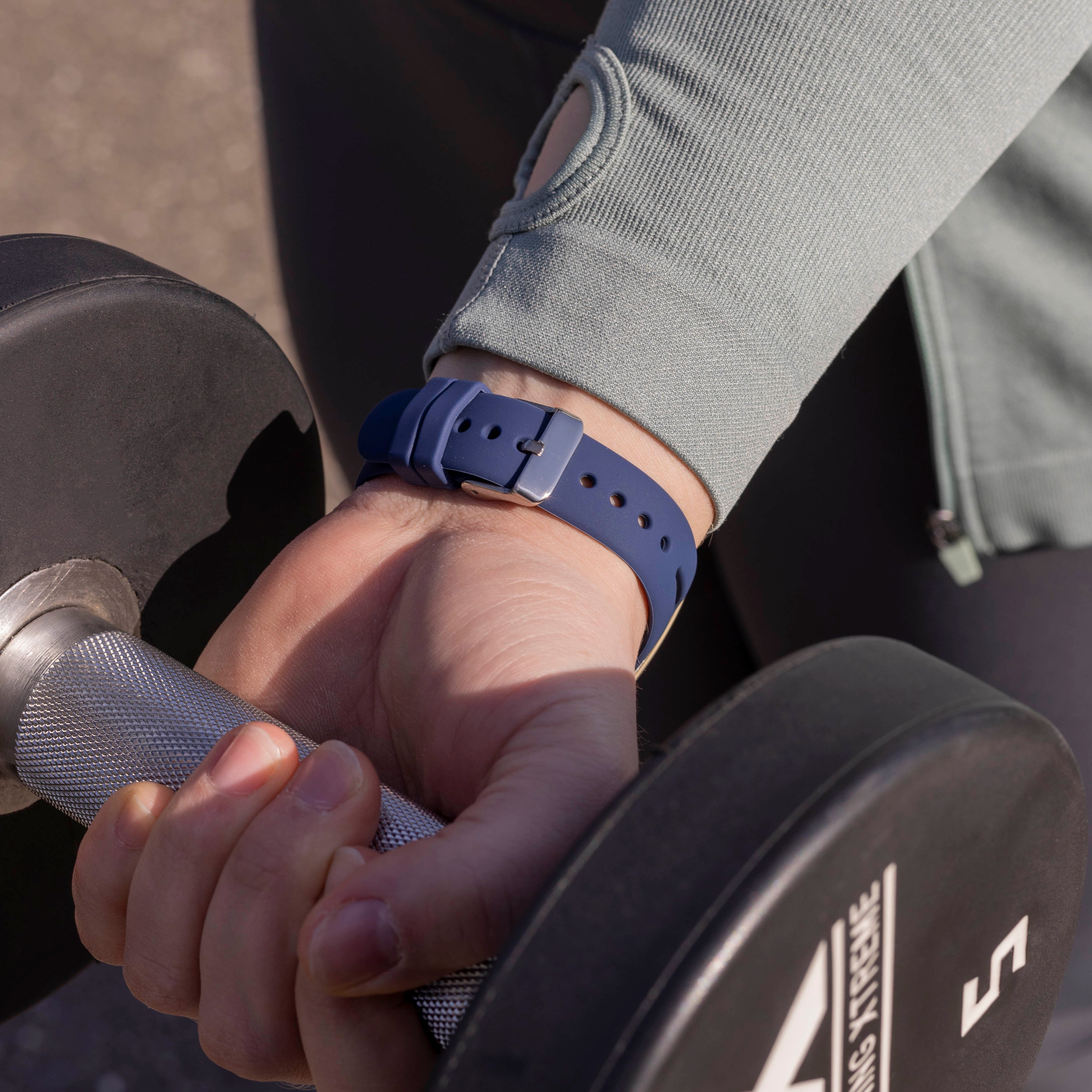 Eine Hand hält eine Hantel. Am Handgelenk befindet sich eine blaue Uhr. Die Person trägt eine graue Jacke. Der Hintergrund ist verschwommen.