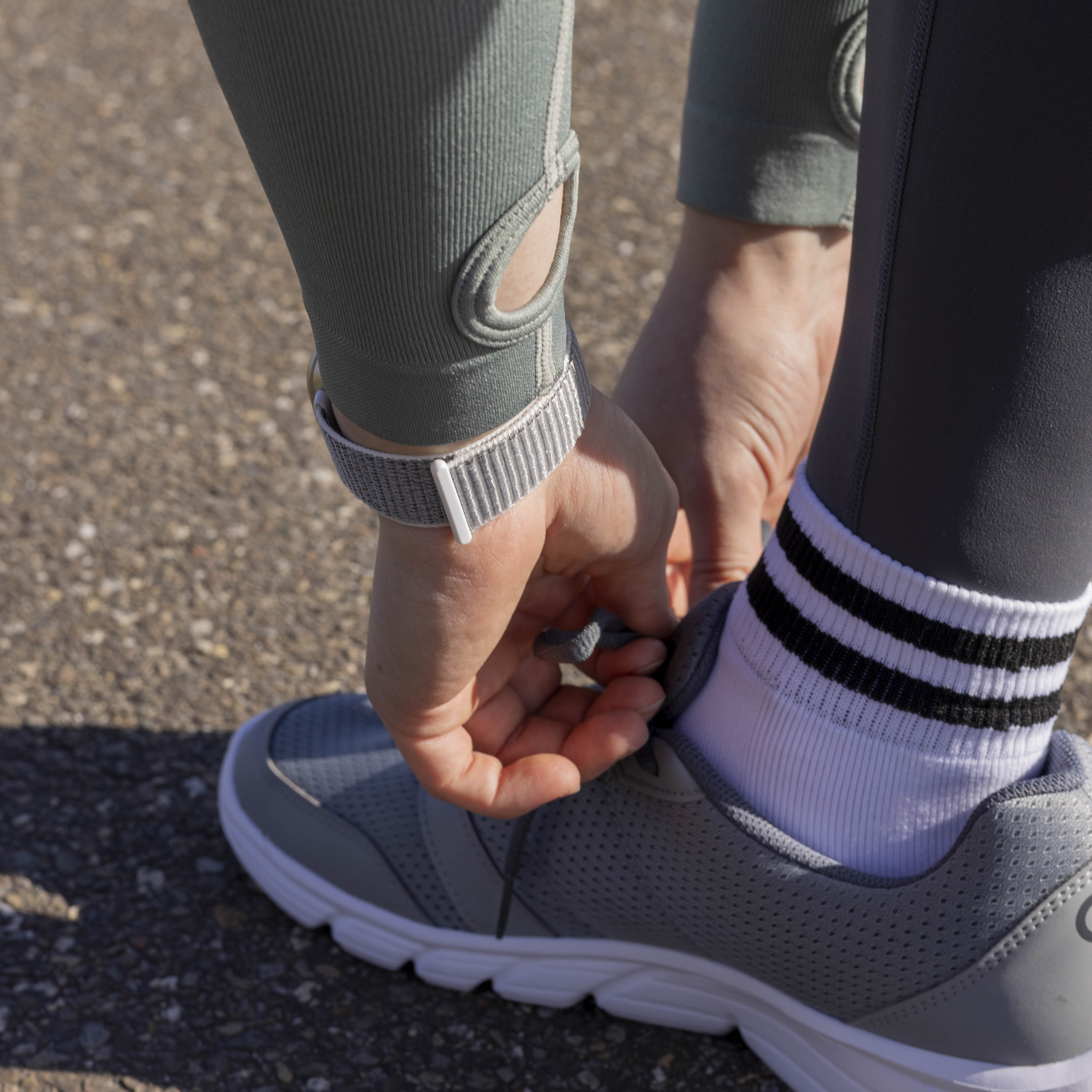 Person bindet Schnürsenkel an einem grauen Sneaker. Hände, eine Uhr und Sportkleidung sind sichtbar. Der Hintergrund ist Asphalt.