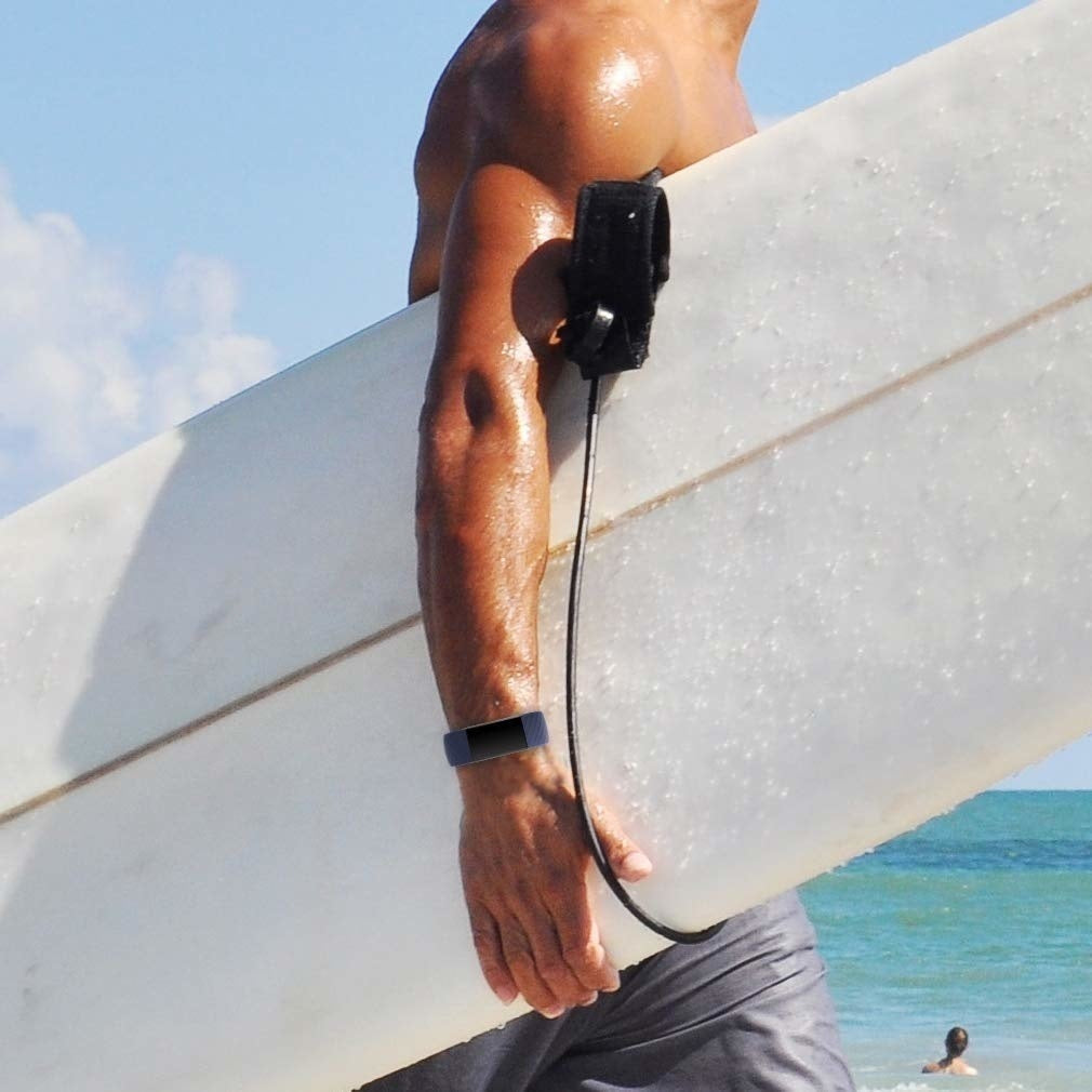 Ein Mann mit Surfbrett steht am Strand. Er hält das Board unter dem Arm. Im Hintergrund ist das Meer und der Himmel ist blau.