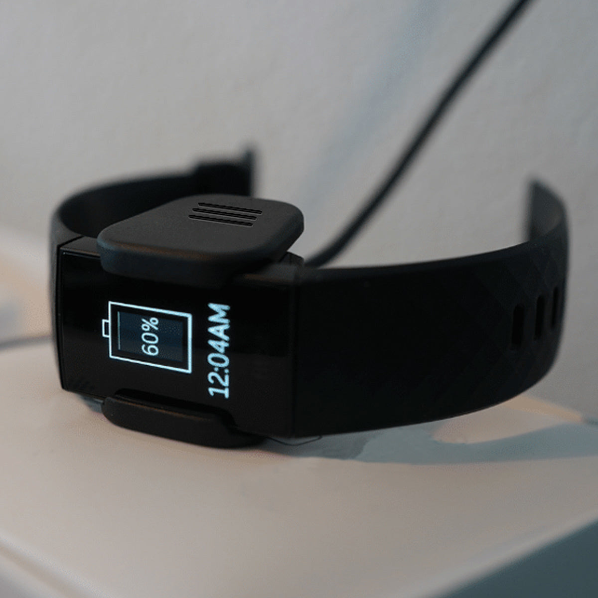 Schwarze Smartwatch wird geladen. Bildschirm zeigt 60 % Akku und 12:04 Uhr an. Ladekabel angeschlossen. Weißer Oberflächenhintergrund.