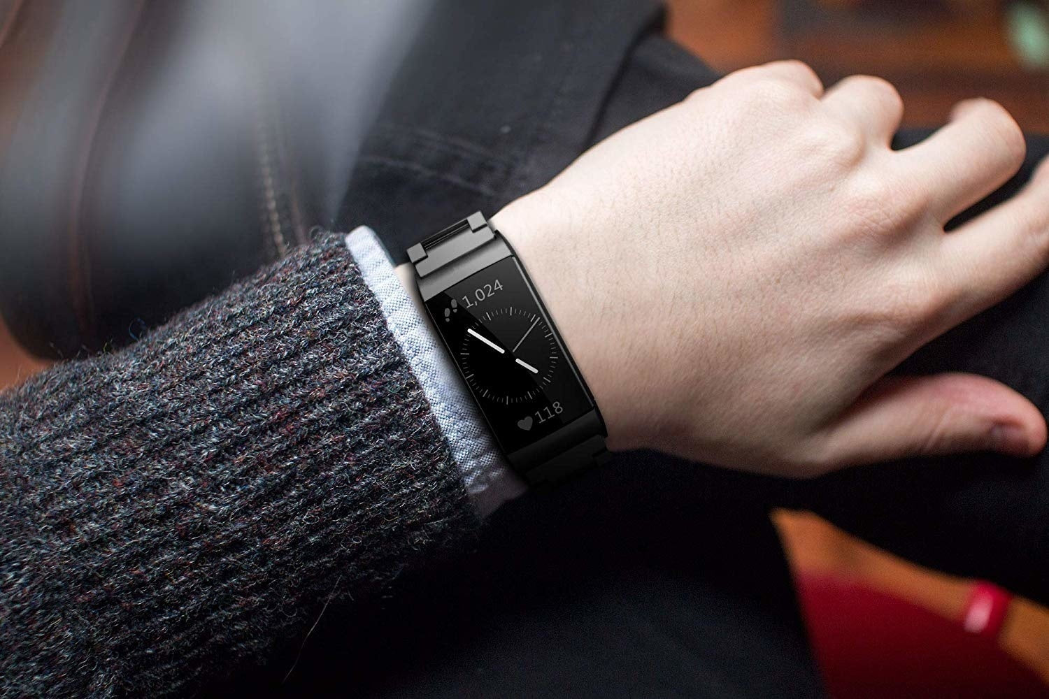 Handgelenk mit schwarzer Smartwatch, die Schritte und Herzfrequenz anzeigt. Die Person trägt einen Pullover und ein Hemd.