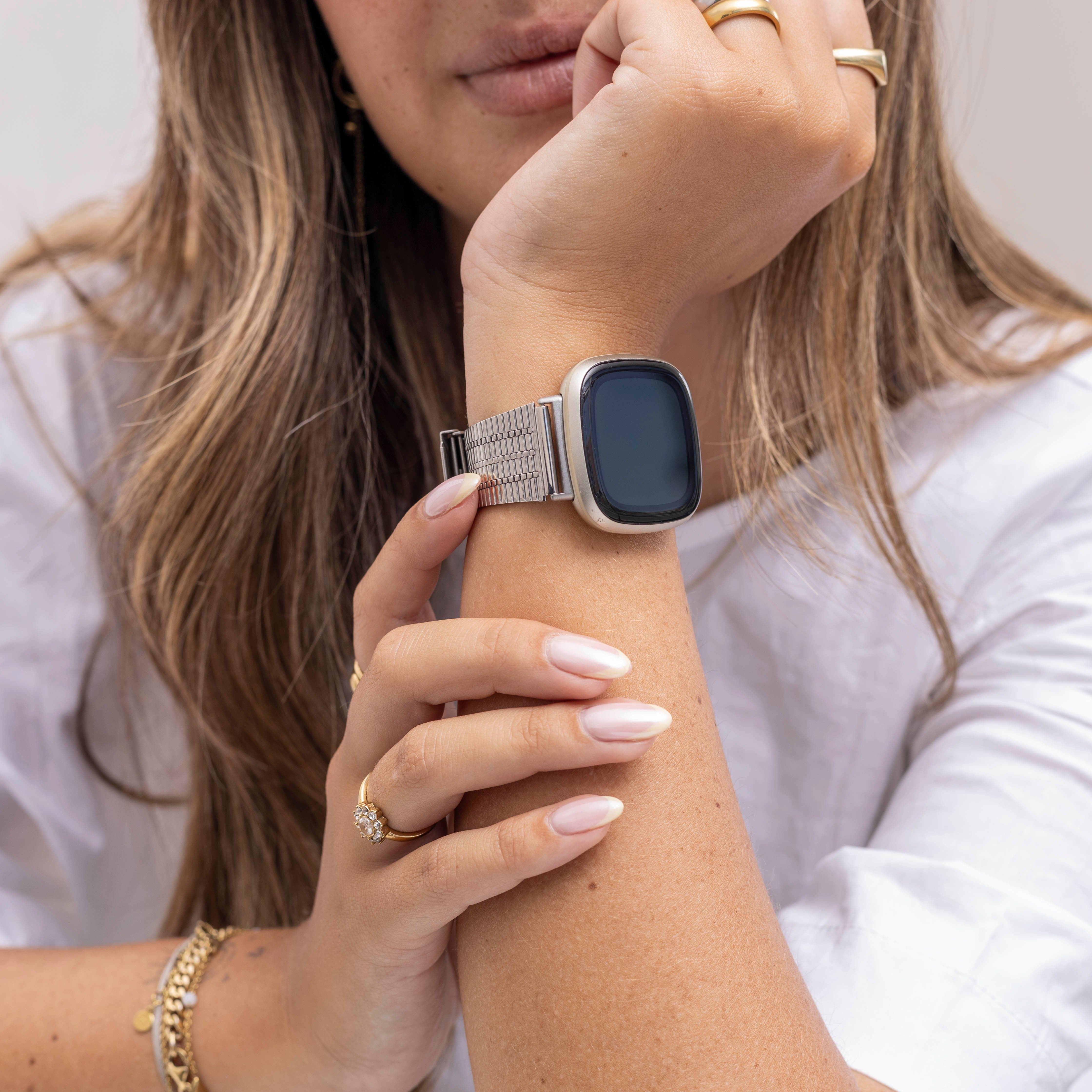 Eine Person, die eine silberne Uhr mit schwarzem Bildschirm, Ringen und einem goldenen Armband trägt. Die Person trägt ein weißes Hemd.