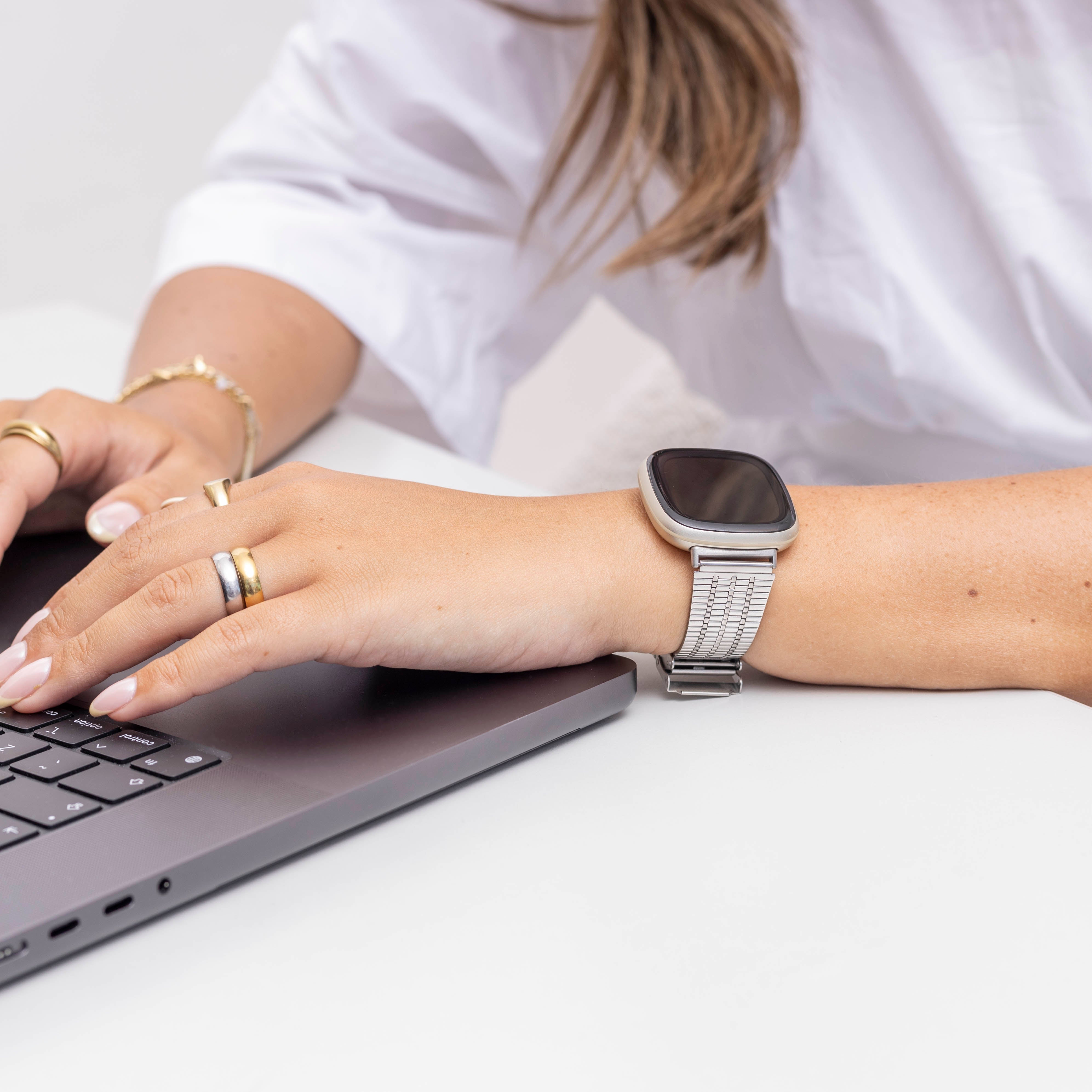 Eine Person, die auf einem Laptop tippt, eine Smartwatch und Ringe trägt. Weißes Hemd und Hintergrund.