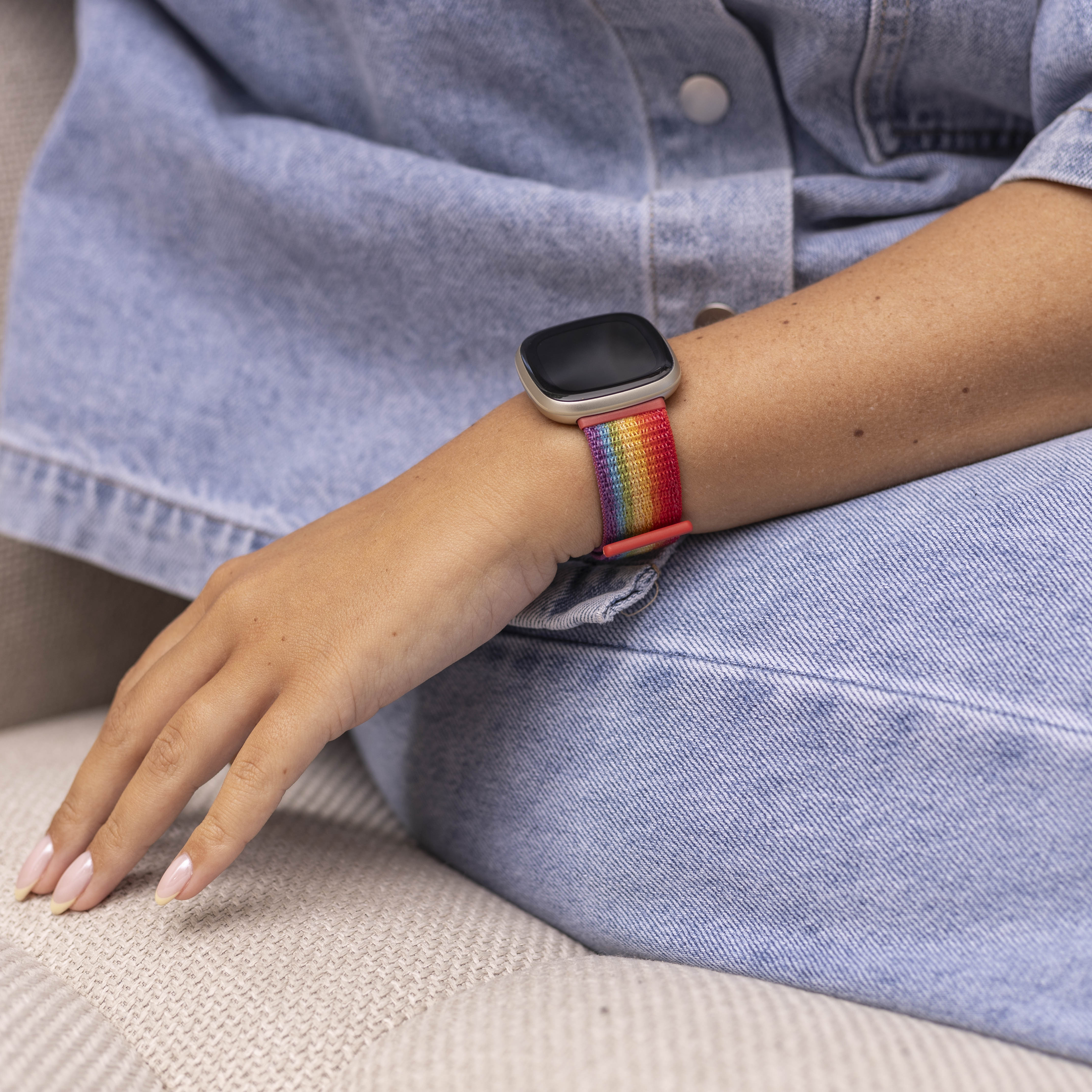 Arm einer Person mit Regenbogen-Armband. Sie trägt eine Jeansjacke und sitzt auf einer beigen Oberfläche.