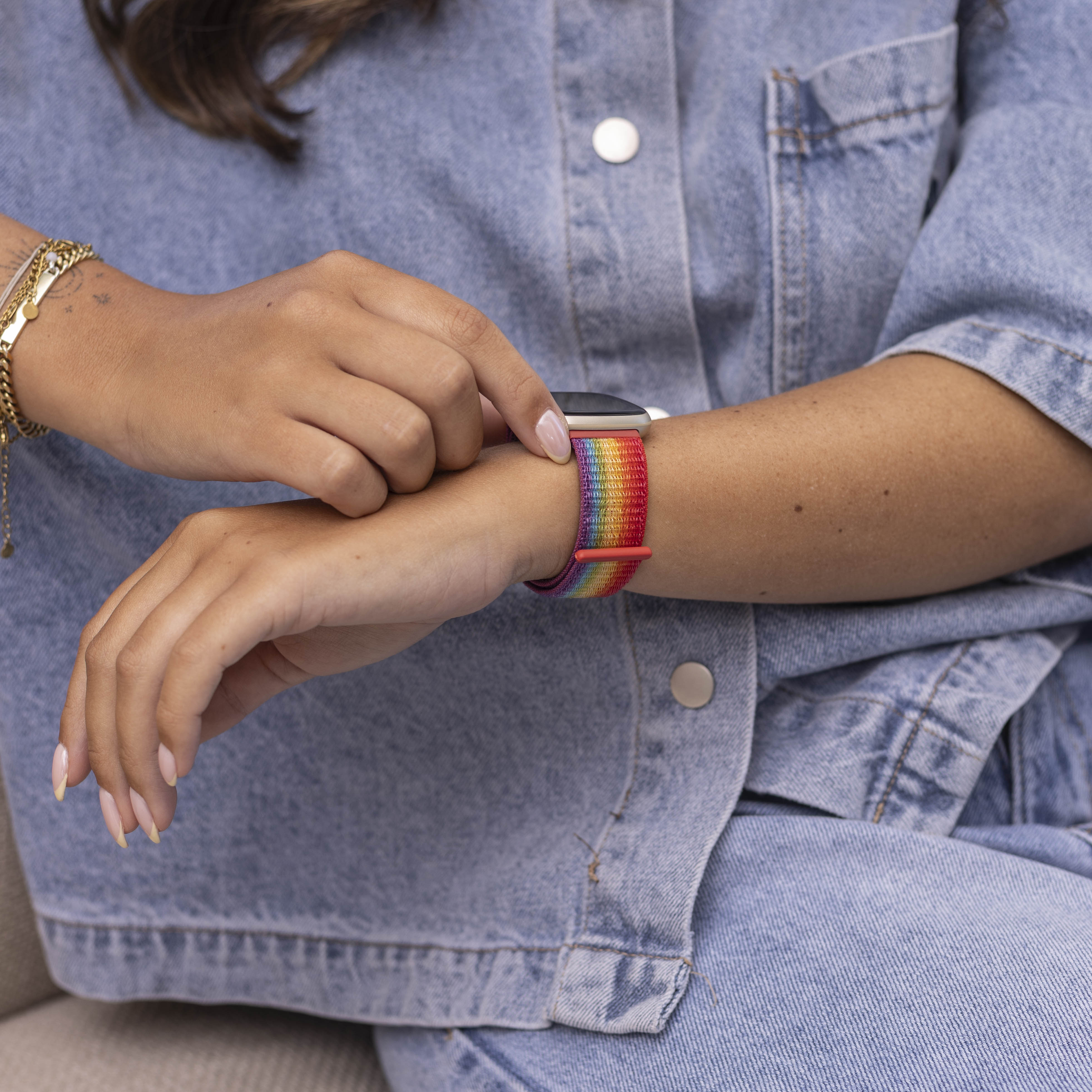 Eine Person mit Jeanshemd und Regenbogen-Armband berührt die Uhr. Goldarmbänder und Tattoos sind sichtbar.