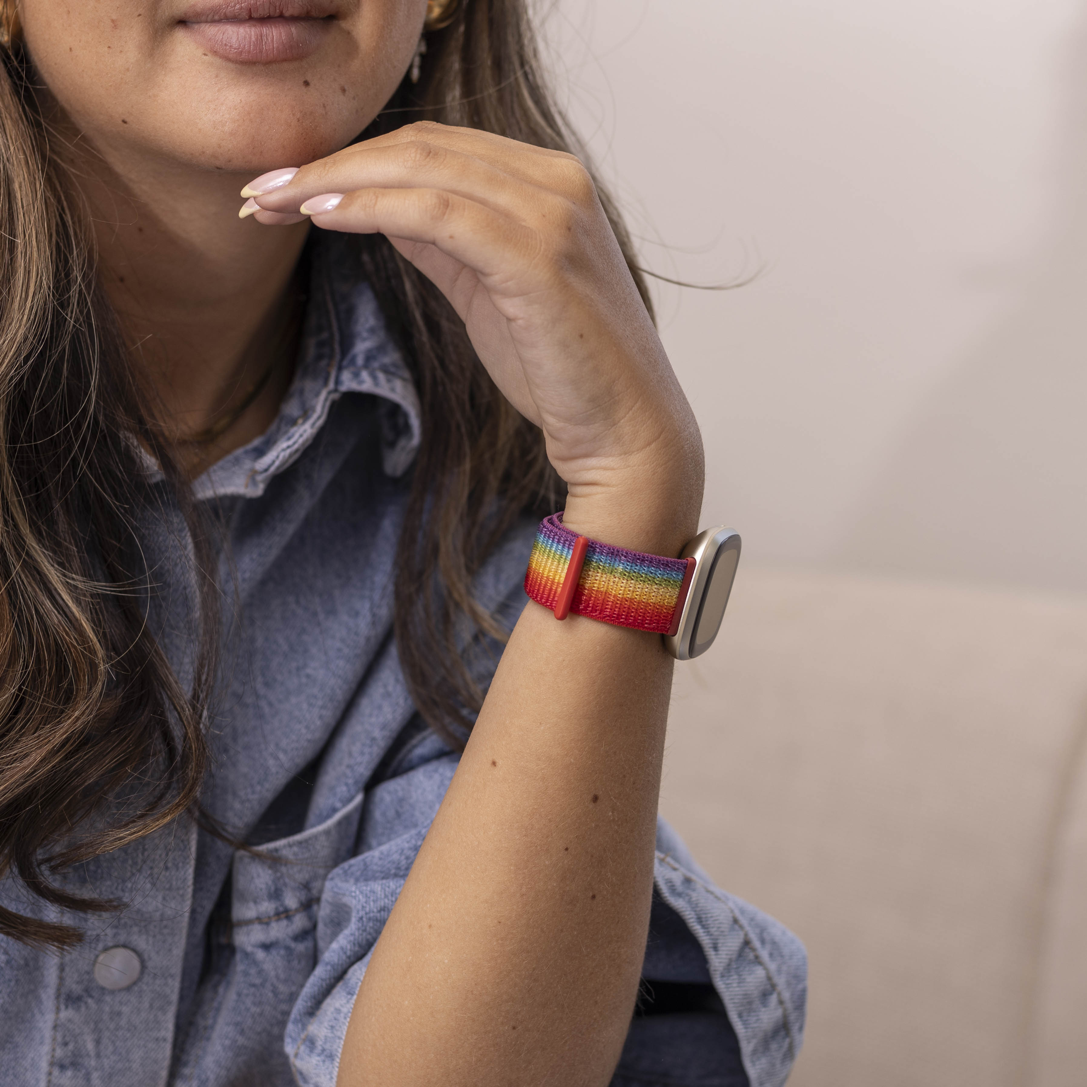 Person mit Regenbogen-Armbanduhr am Handgelenk. Sie trägt eine Jeansjacke und stützt das Kinn auf die Hand.