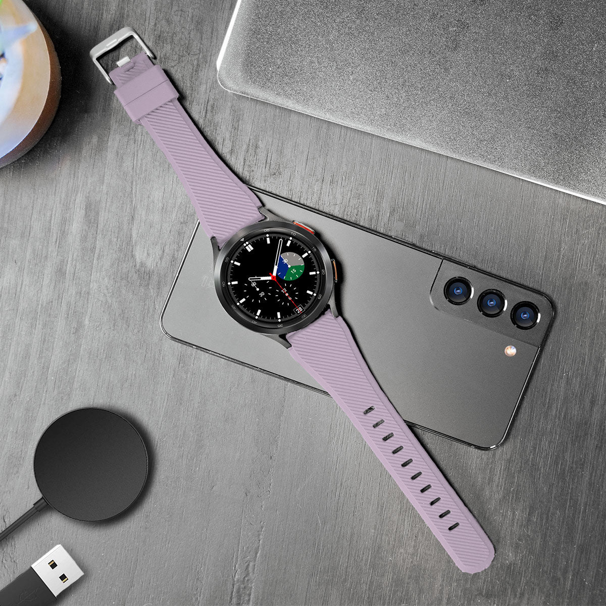 Eine lavendelfarbene Smartwatch auf einem Telefon und einer Holzoberfläche. Ein Ladepad und ein Laptop sind ebenfalls vorhanden.