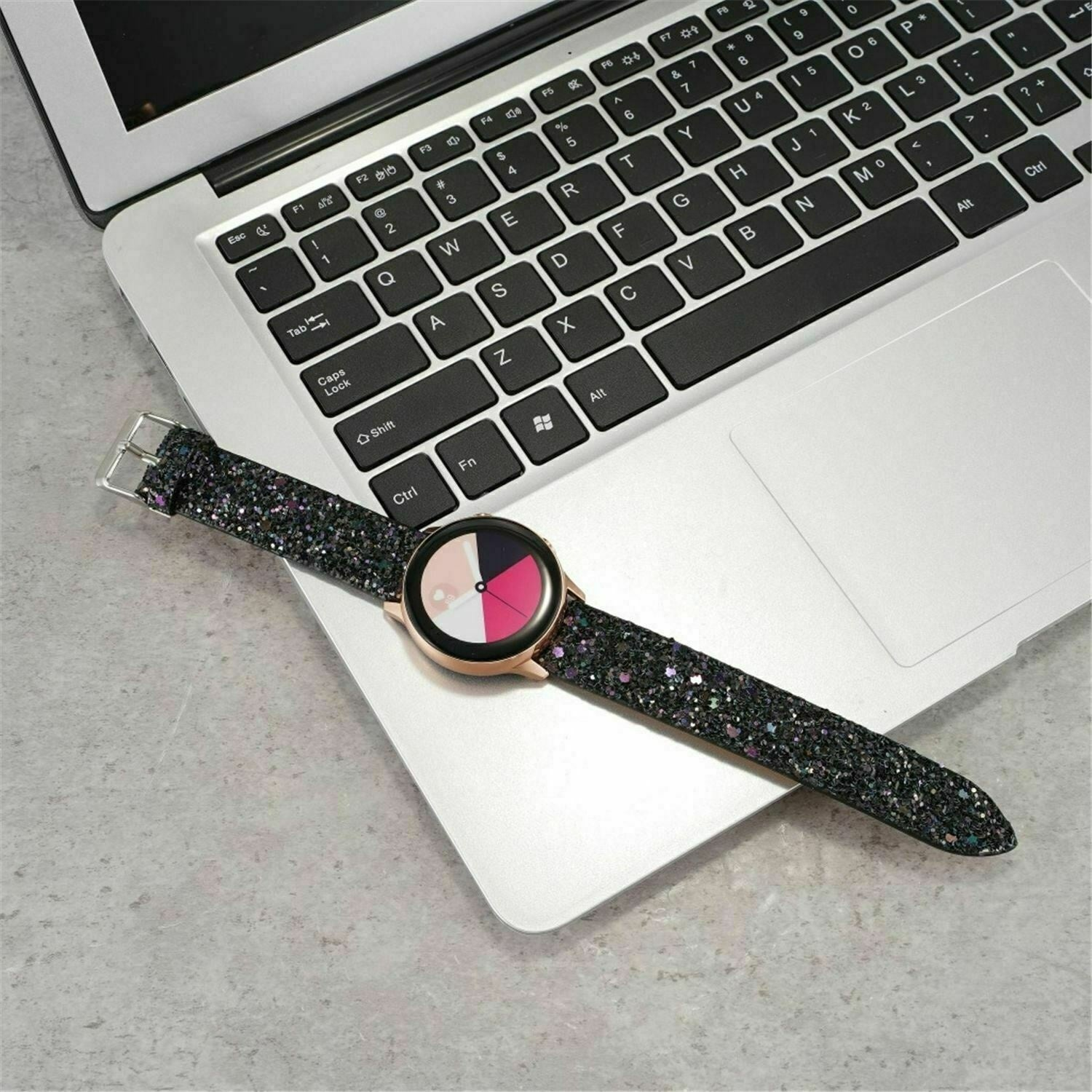 Eine Smartwatch mit glitzerndem Armband liegt auf einem Laptop. Das Zifferblatt zeigt rosa, weiße und schwarze Segmente. Der Laptop ist geöffnet.