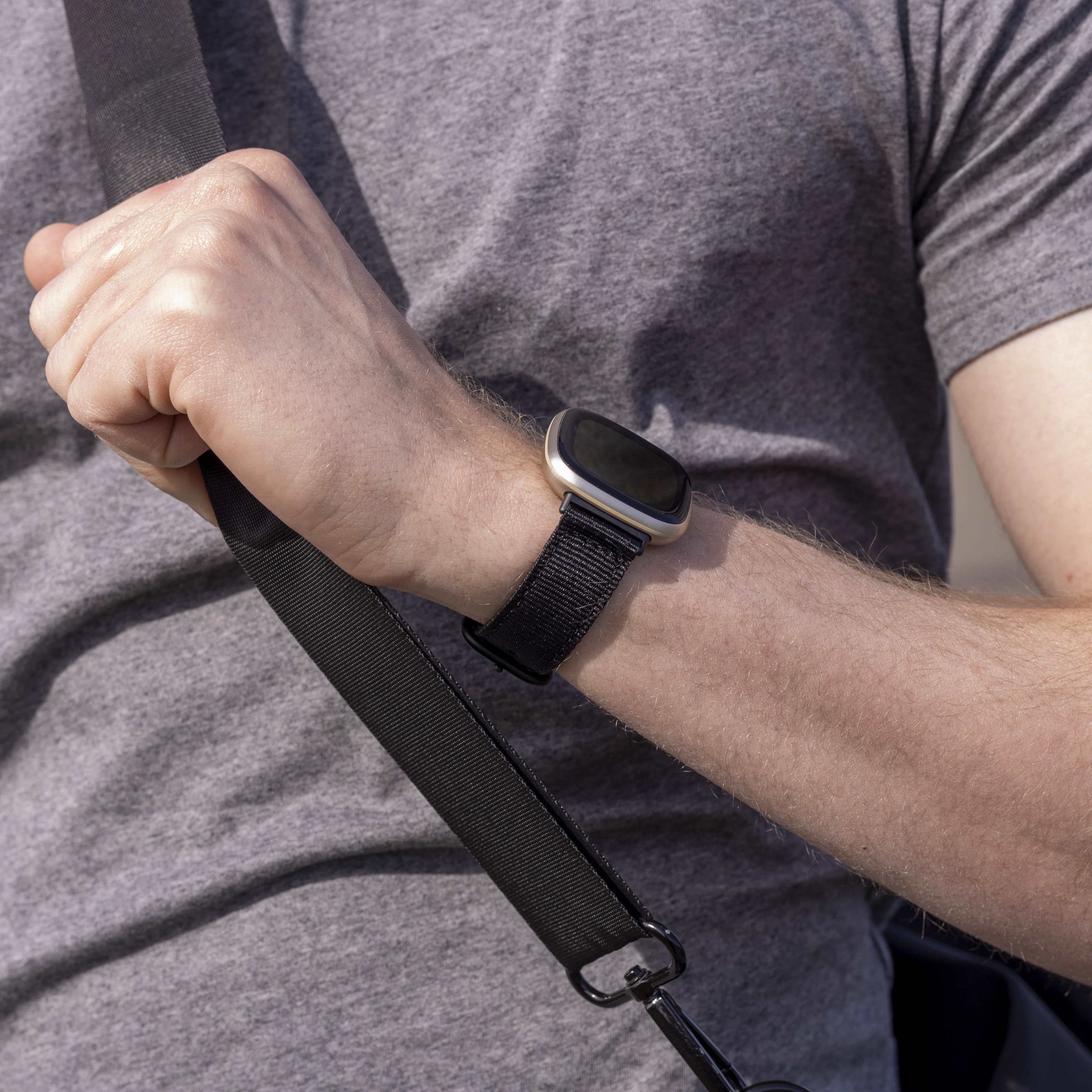 Eine Person hält einen schwarzen Taschenriemen. Eine goldene Smartwatch befindet sich an ihrem Handgelenk. Sie tragen ein graues Hemd.